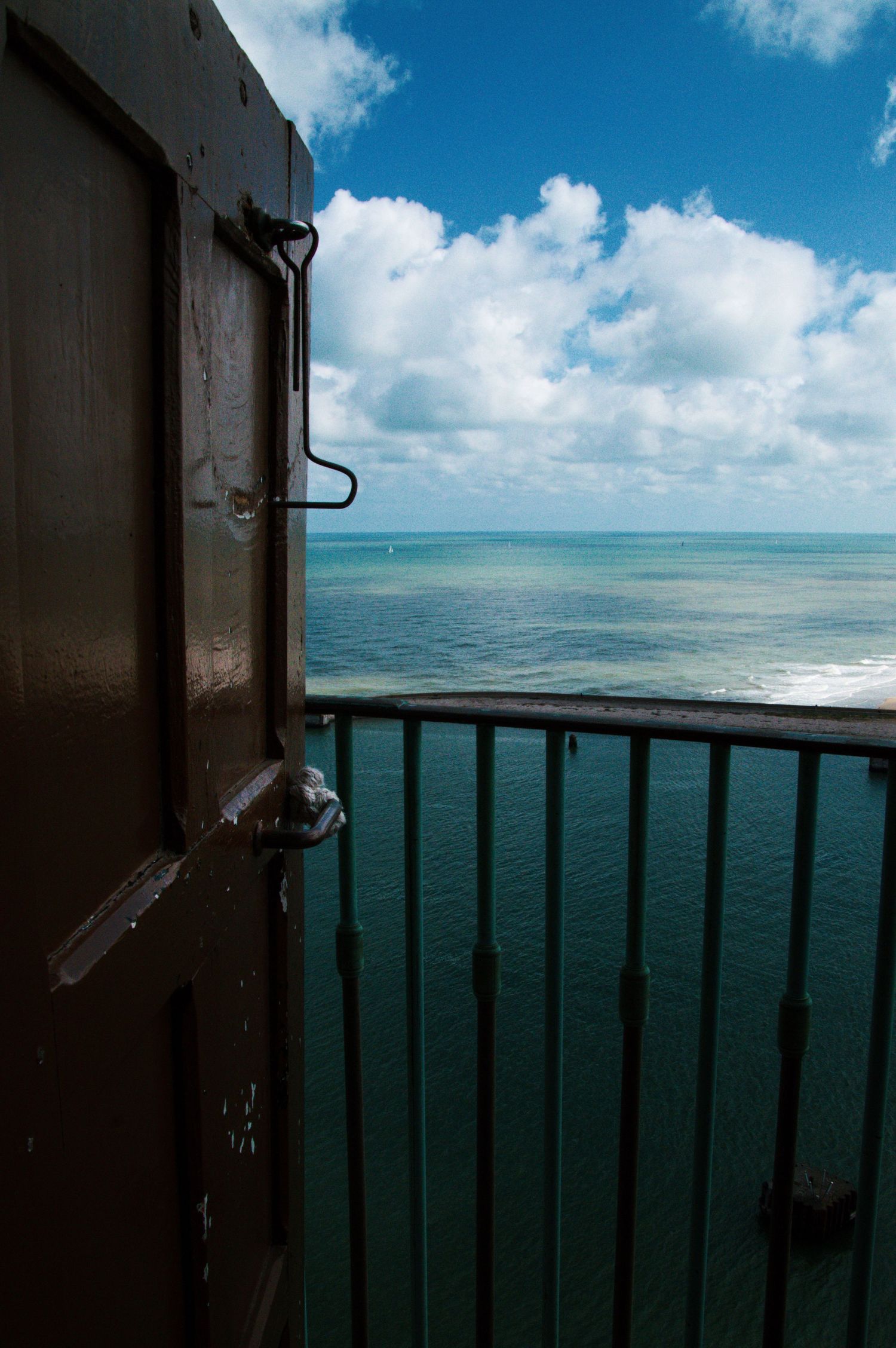 Open metal door leading to a balcony with iron railings overlooking a turquoise ocean under a blue cloudy sky.