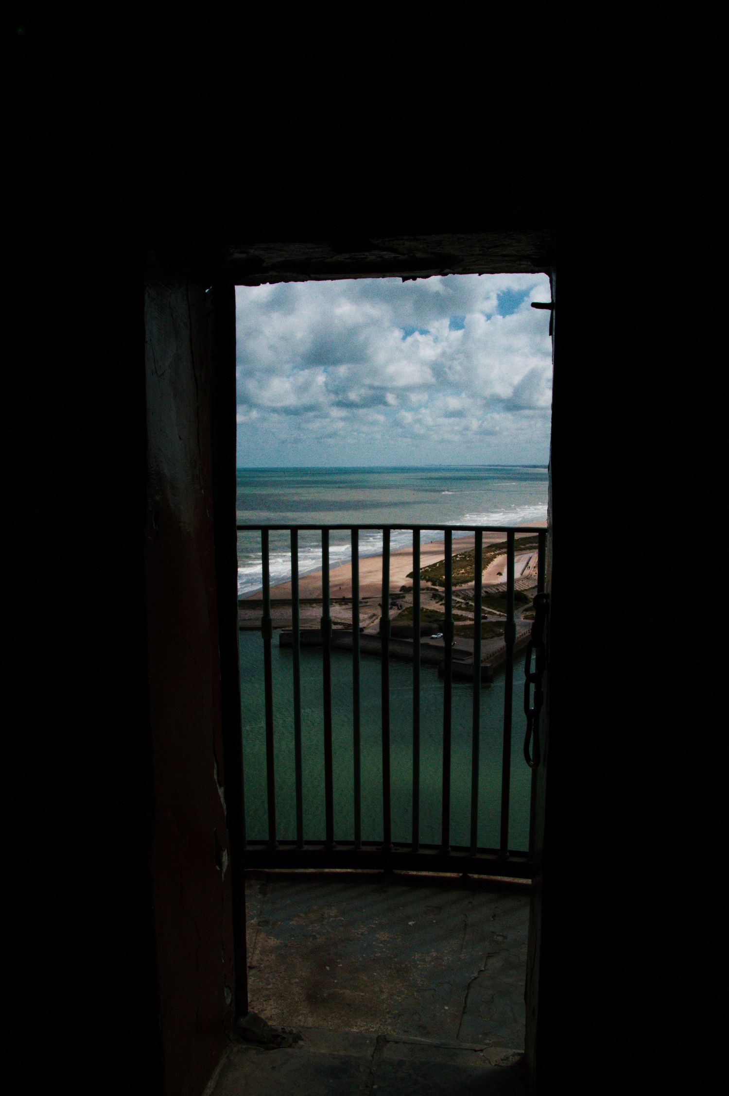 Dark doorway opens to a balcony with iron railing overlooking turquoise ocean waters and cloudy sky.