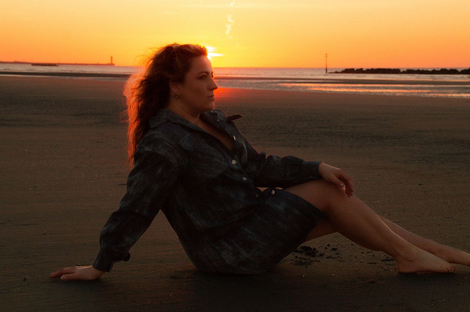 Woman with red hair sitting on a beach at sunset, wearing a dark jacket, with golden sky reflecting on wet sand.