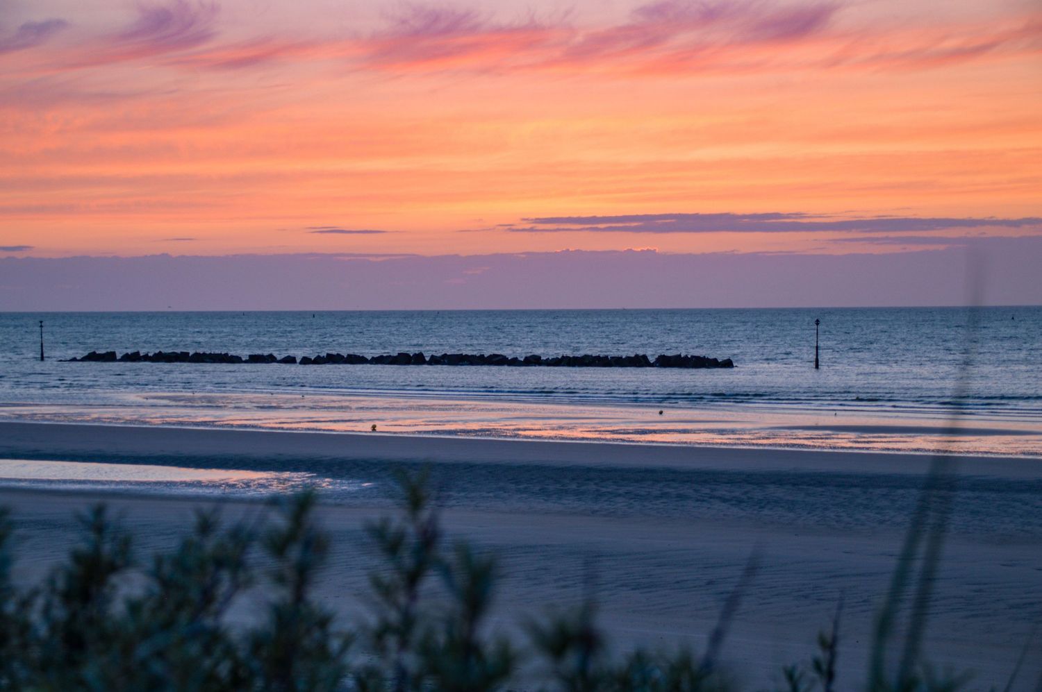 Vibrant orange and pink sunset over a calm beach with silhouetted breakwater in the distance.
