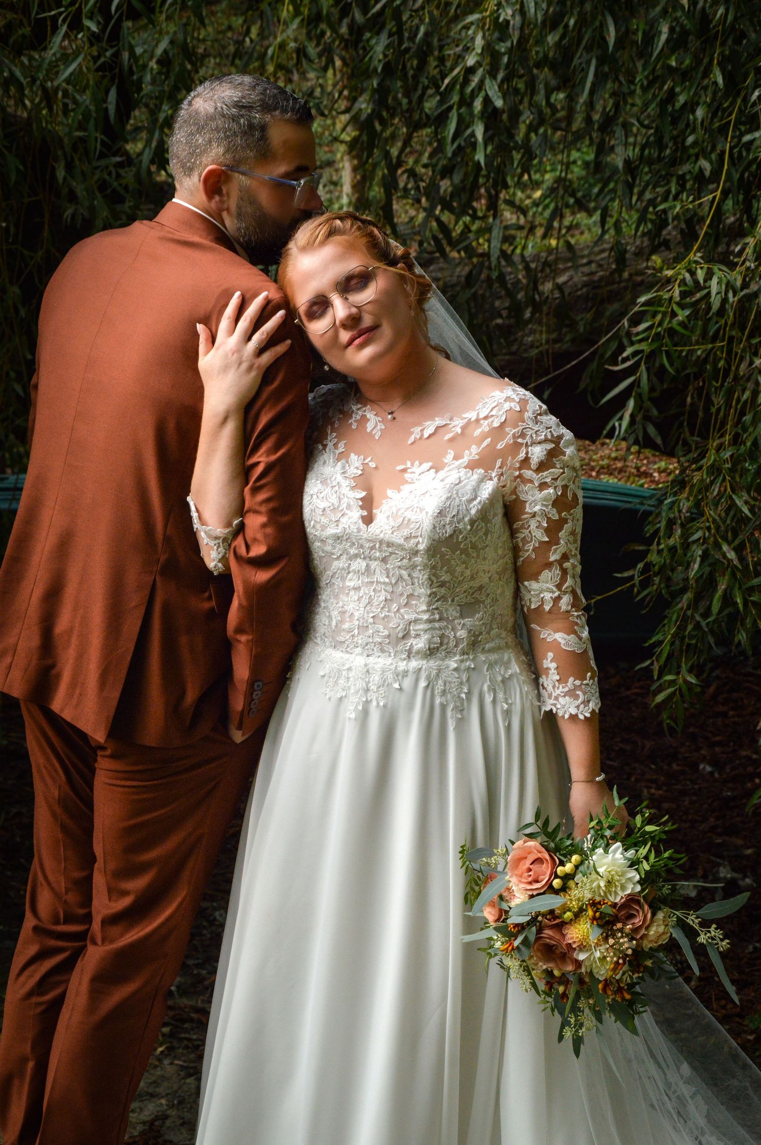 Romantic wedding portraits under willow trees with bride in white lace dress and groom in brown suit.