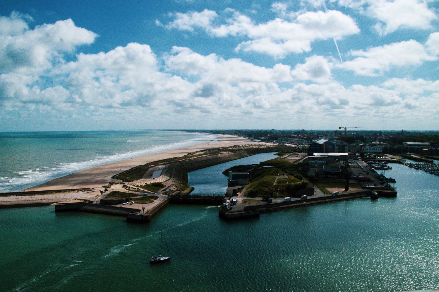 Aerial view of a coastal harbor with sandy beaches, calm blue waters, and a small boat near the shore under a cloudy sky.