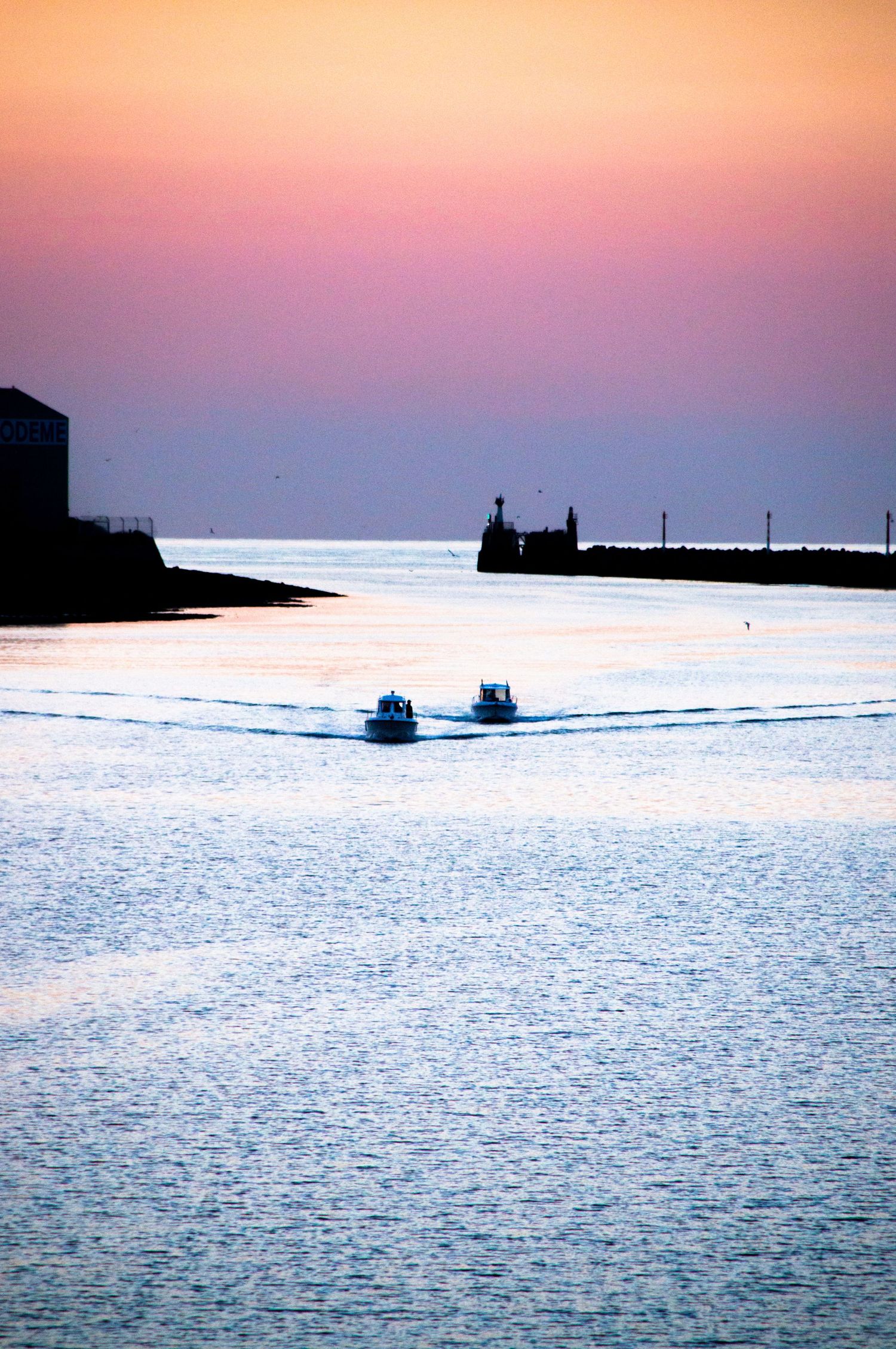 Two boats sailing into a harbor at sunset with vibrant orange and purple sky reflecting on calm blue water.