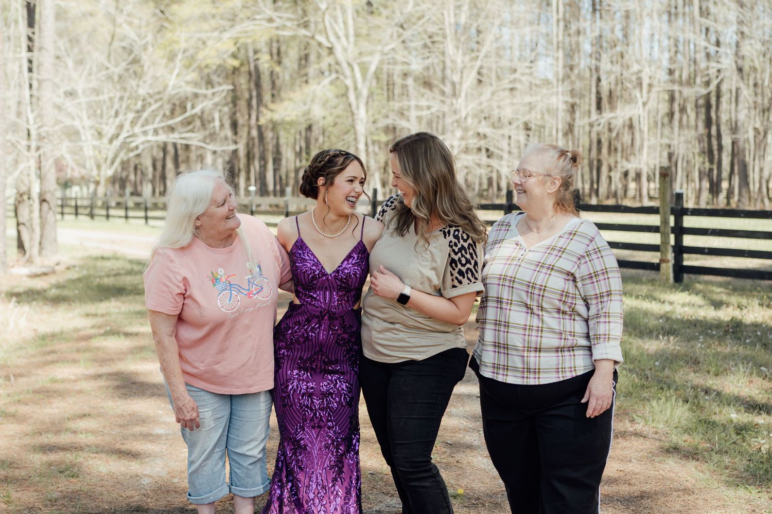 Four women laughing and walking together outdoors, one wearing a purple sequin dress, surrounded by trees.