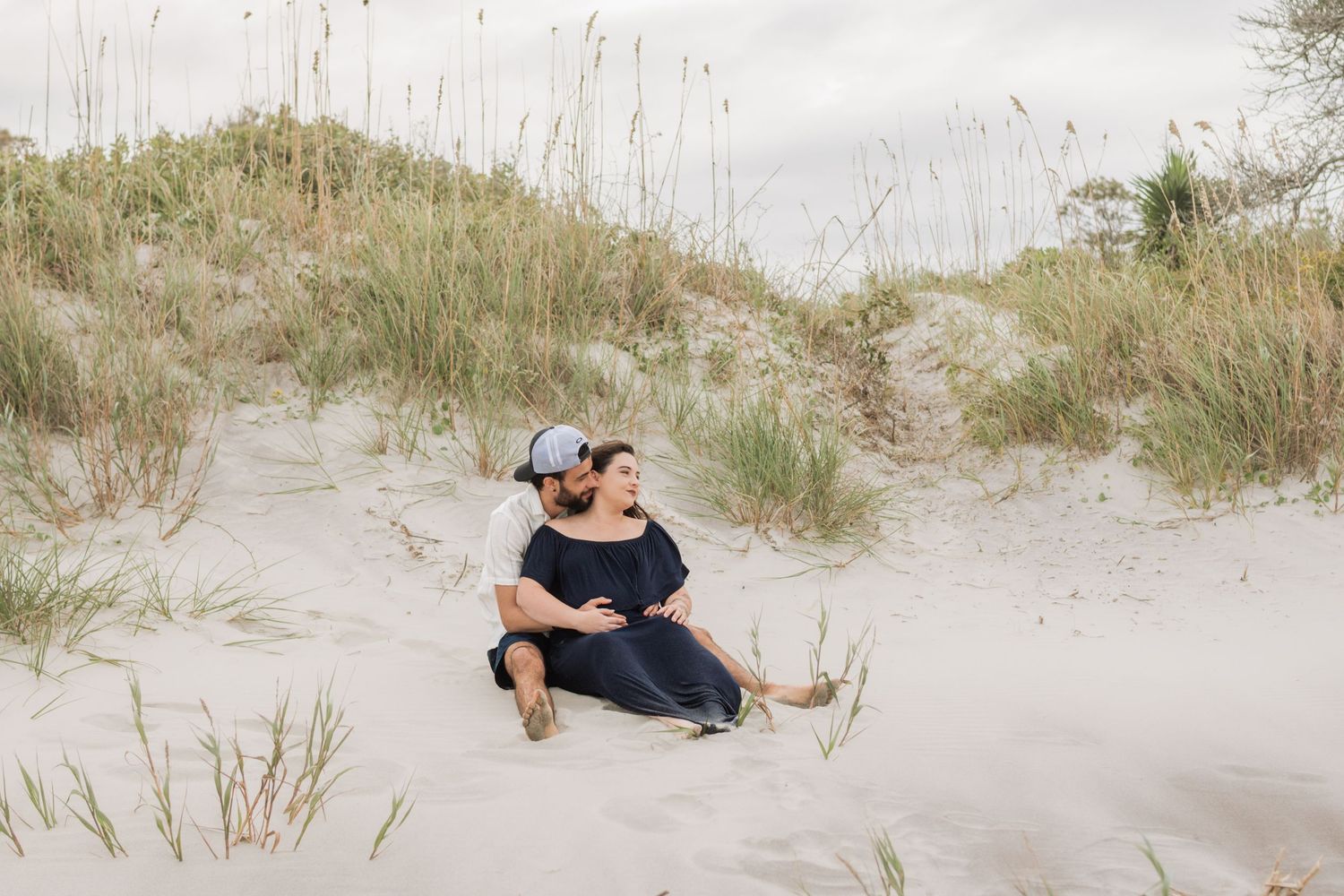 Couple sitting together on sandy beach dunes surrounded by coastal grass during a maternity photo shoot.