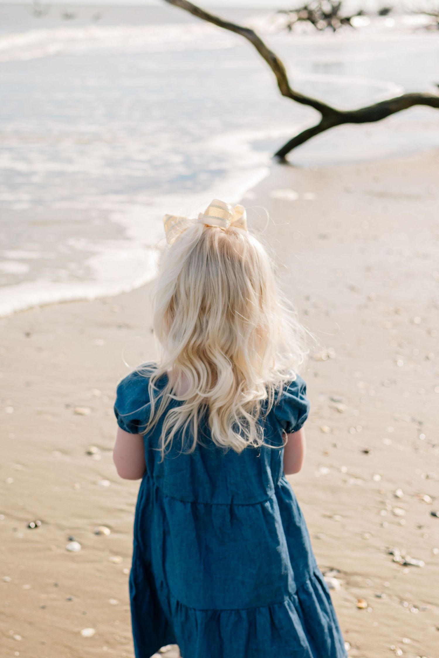 A young blonde girl in a teal dress with a bow stands on a sandy beach facing the ocean waves.