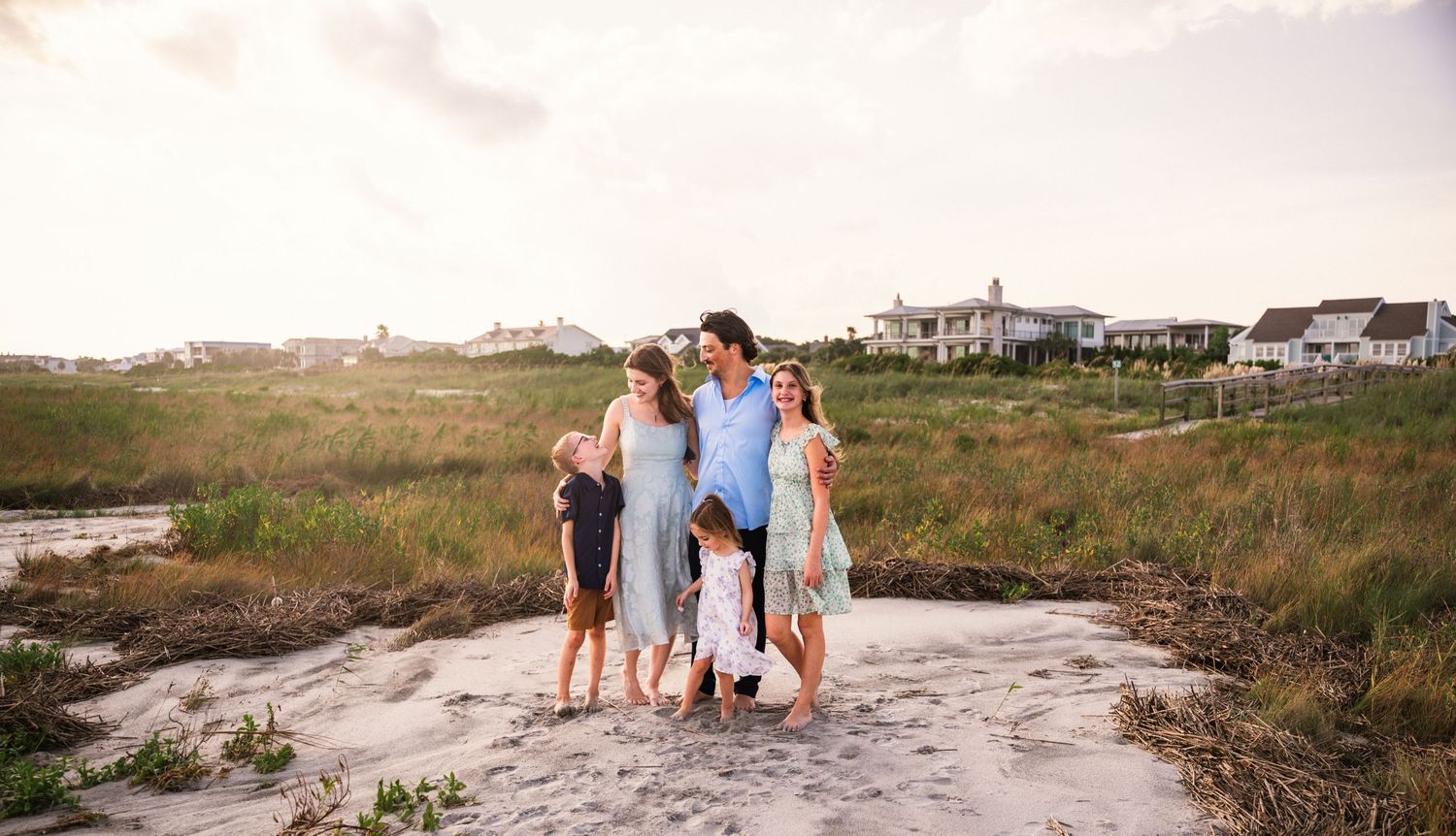 Family of five posing together on a sandy beach path surrounded by coastal grass and homes at golden hour.