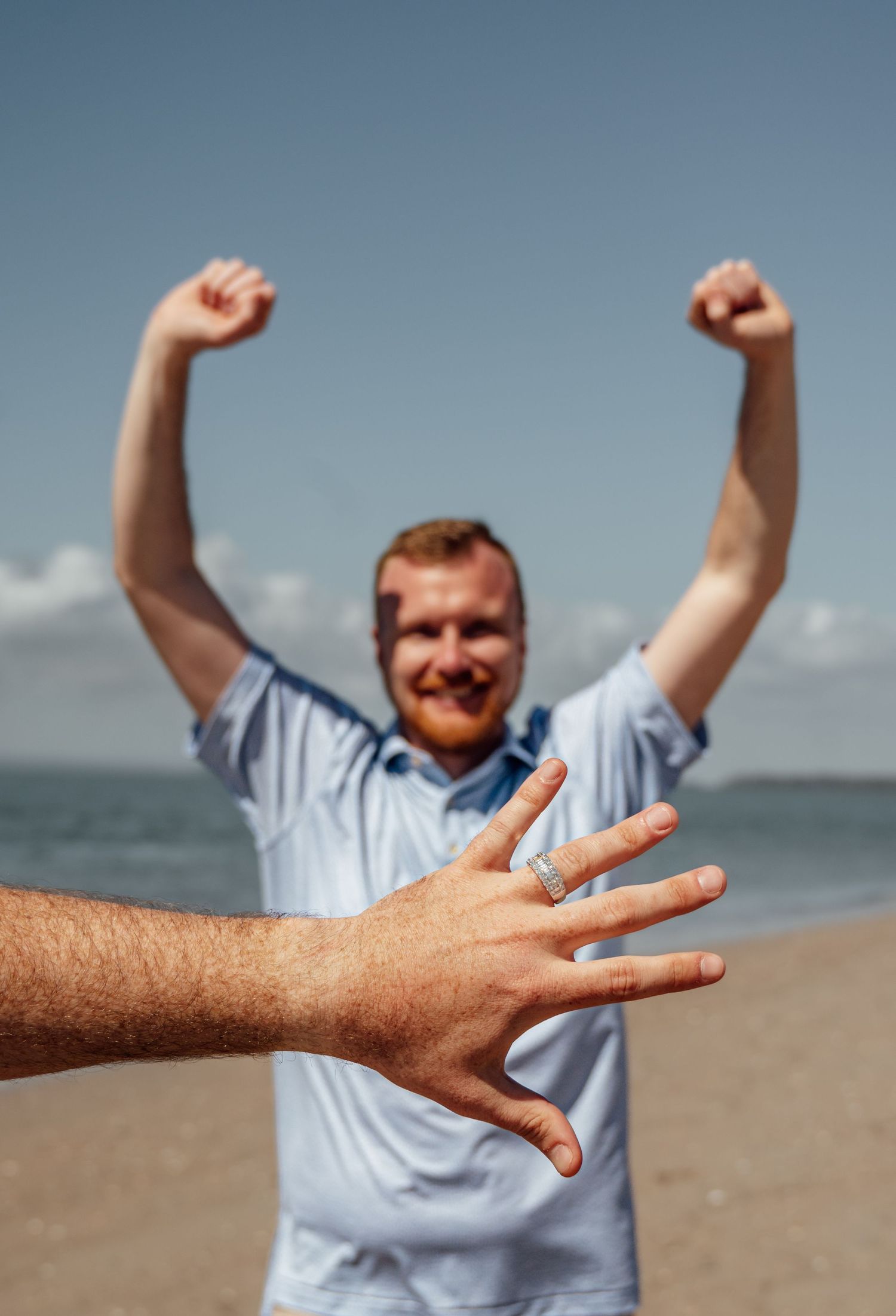 A joyful man raises his fists in celebration on a beach as a hand displays an engagement ring in the foreground.
