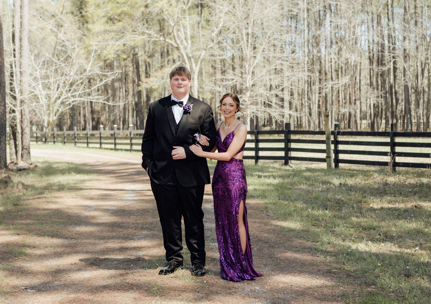 A young couple dressed in prom attire, tuxedo and purple sequin gown, posing on a dirt path near a wooden fence.