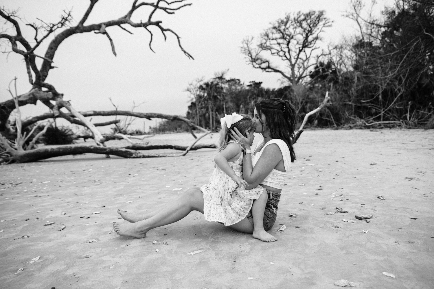 A black and white photo of a woman and young girl sharing a tender moment sitting on a sandy beach.