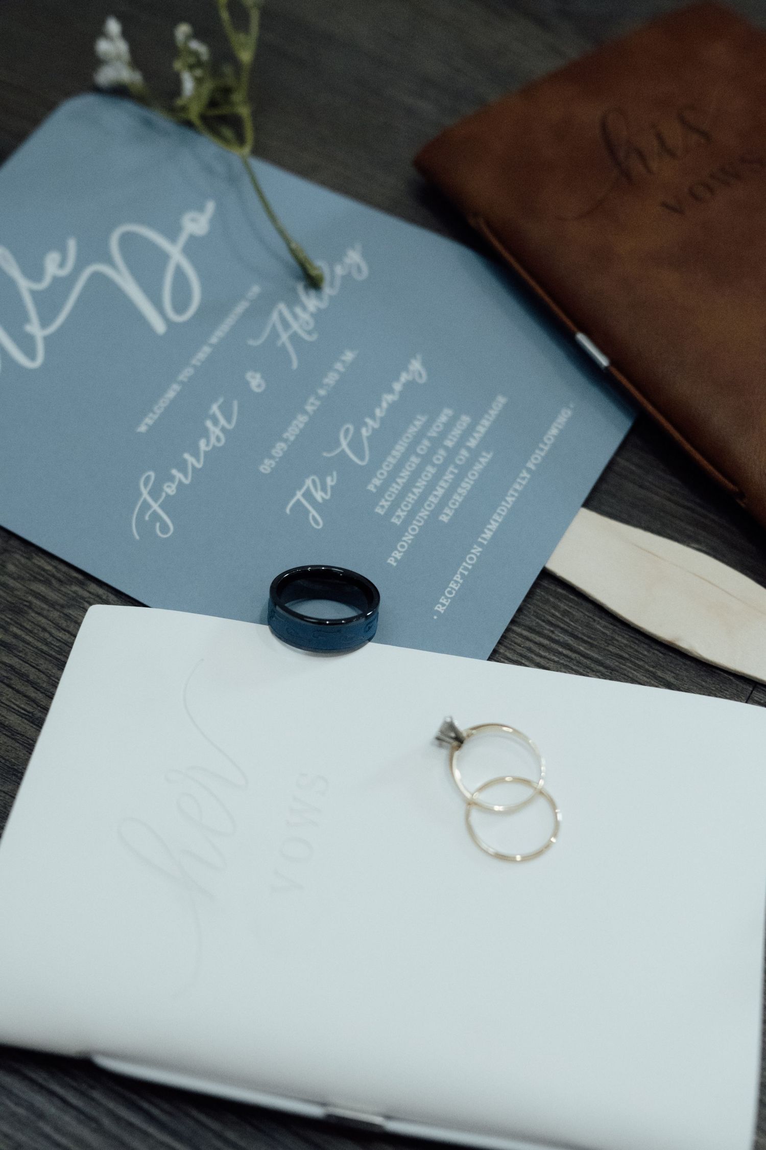 Elegant wedding flat lay with dusty blue invitation, black band ring, diamond rings, and brown leather vow book on dark wood.