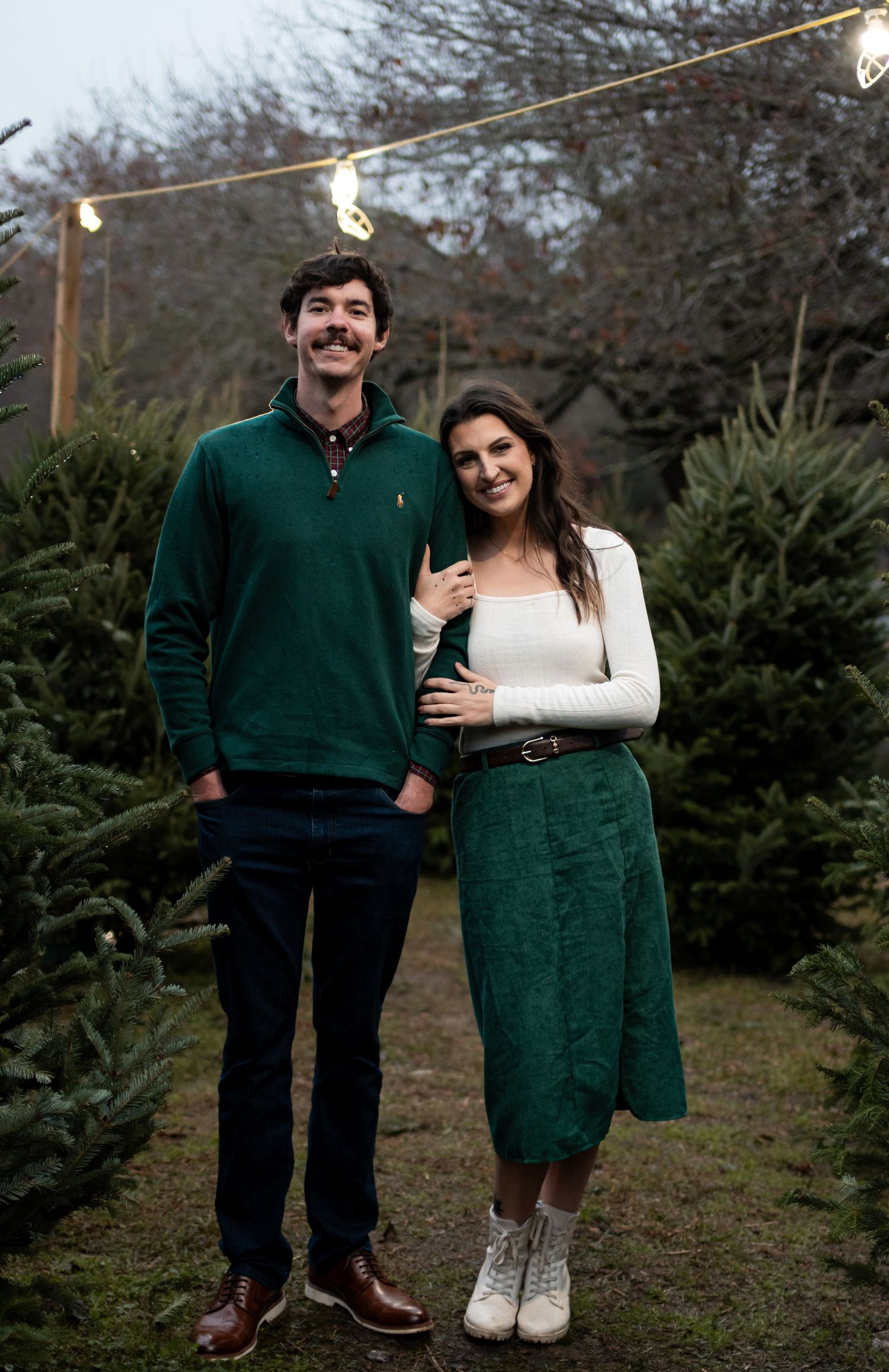 Couple smiling at a Christmas tree farm, man in green sweater and woman in white top with green skirt.