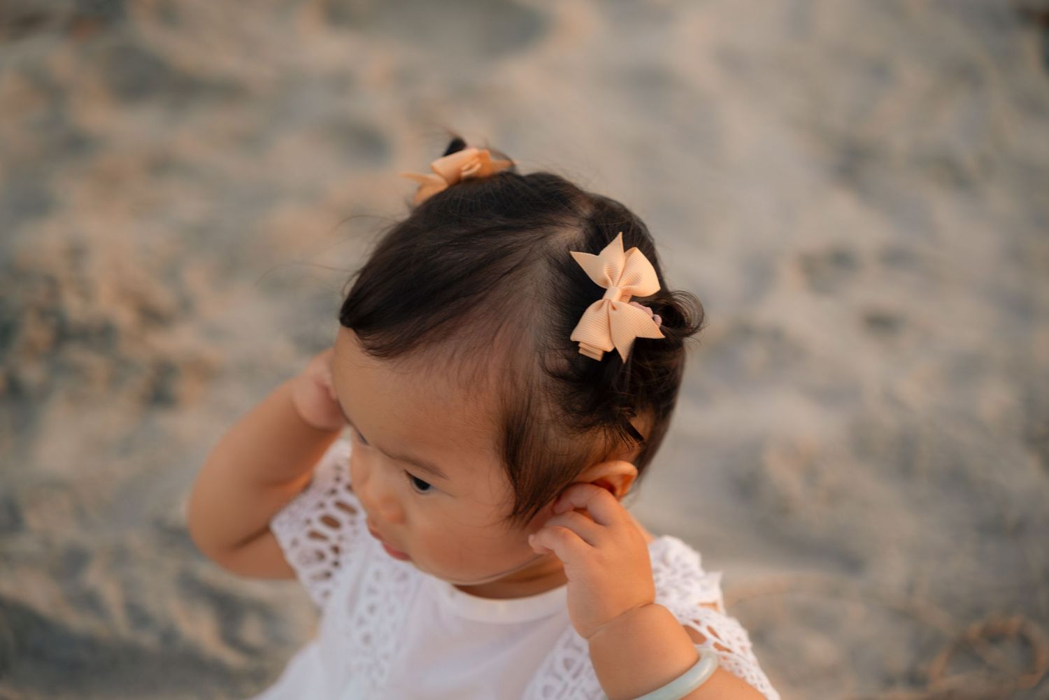 A baby girl with a bow hair clip in dark hair wears a white dress on a sandy beach.