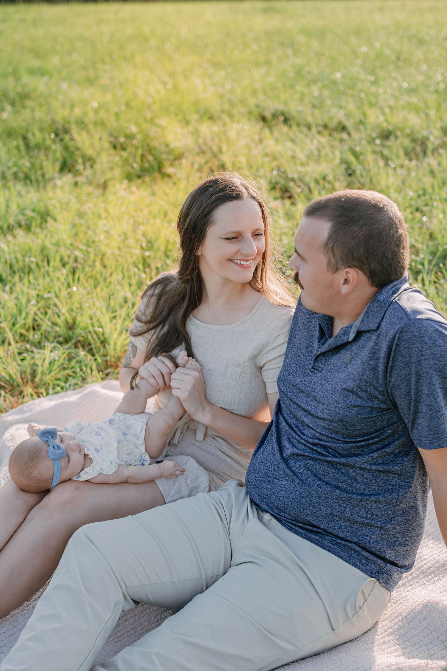 A happy young family sits on a blanket in a green field, with a mother holding her newborn baby while smiling at her husband.