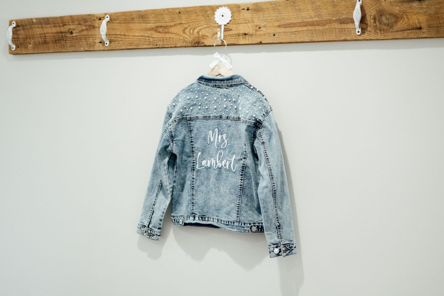 Personalized denim jacket with white cursive text hanging on a wooden coat rack against a white wall.
