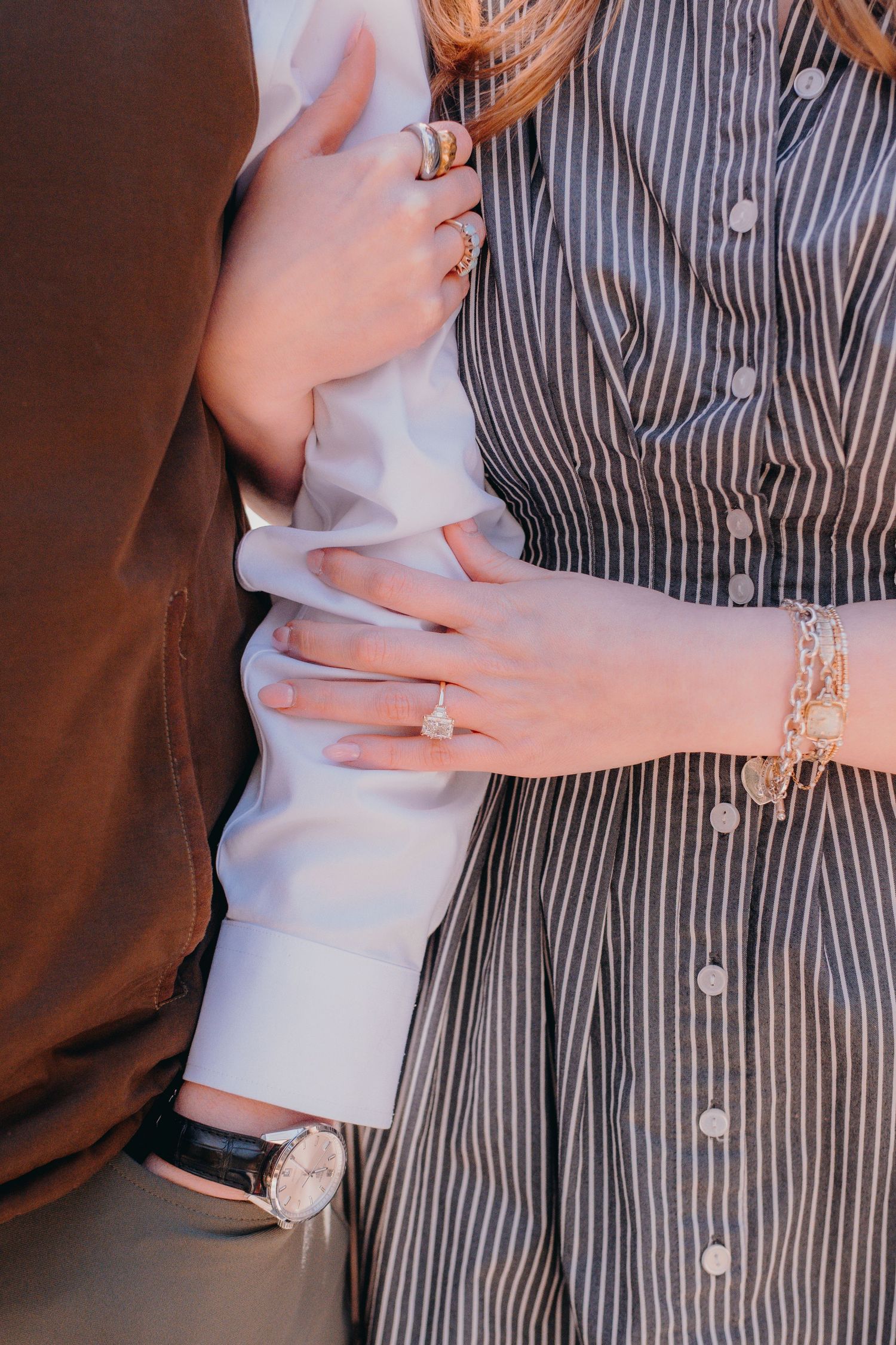 Couple embracing, woman wearing engagement ring, gold bracelet, and rings on striped dress, man in white shirt with watch.