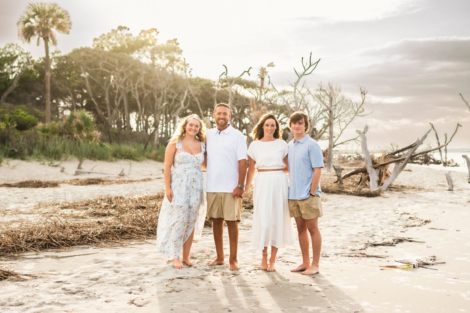 Family of four posing together on a sandy beach with driftwood and palm trees in the background.