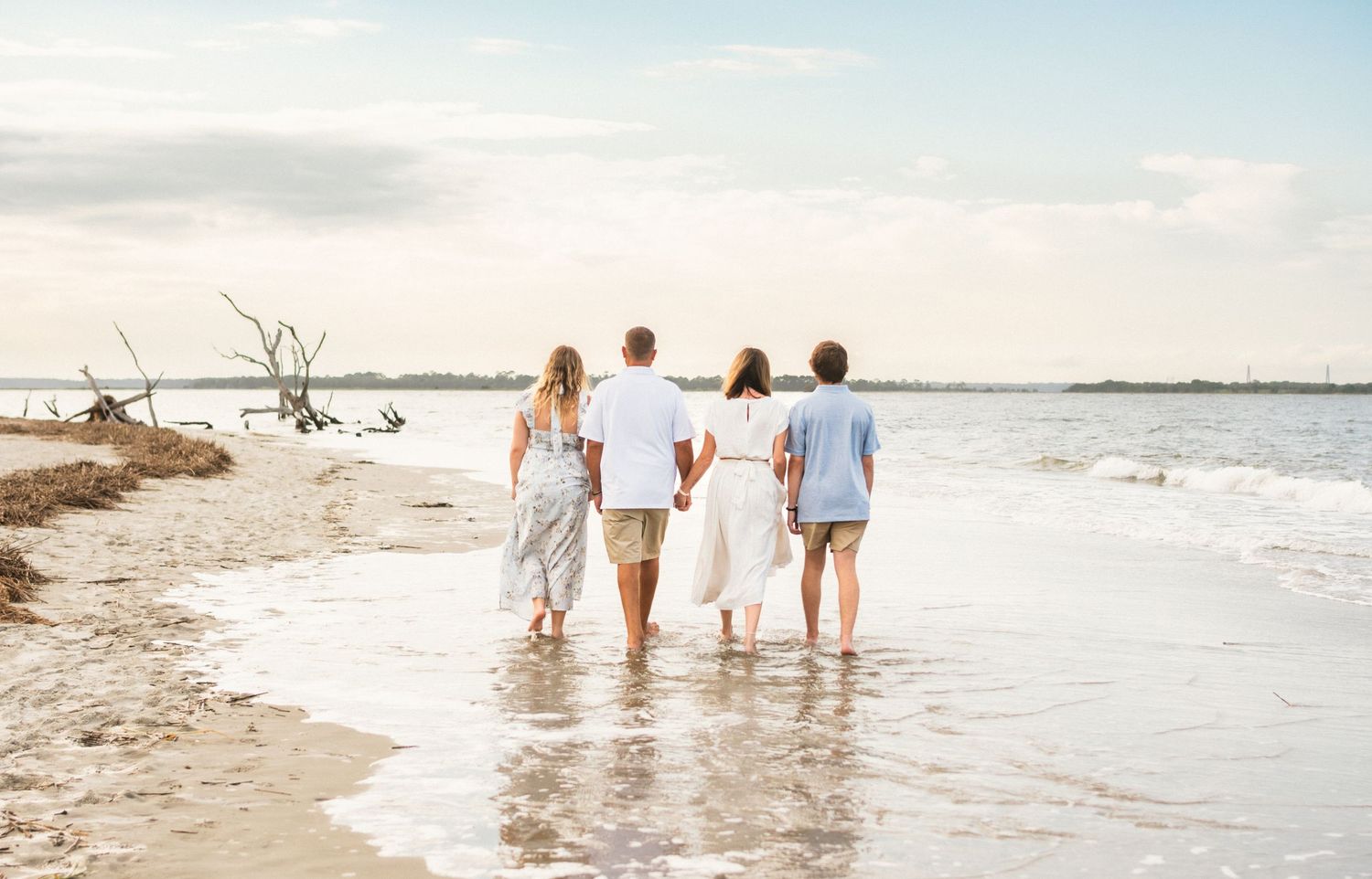 Family of four walking along a sandy beach shoreline together, viewed from behind on a cloudy day.