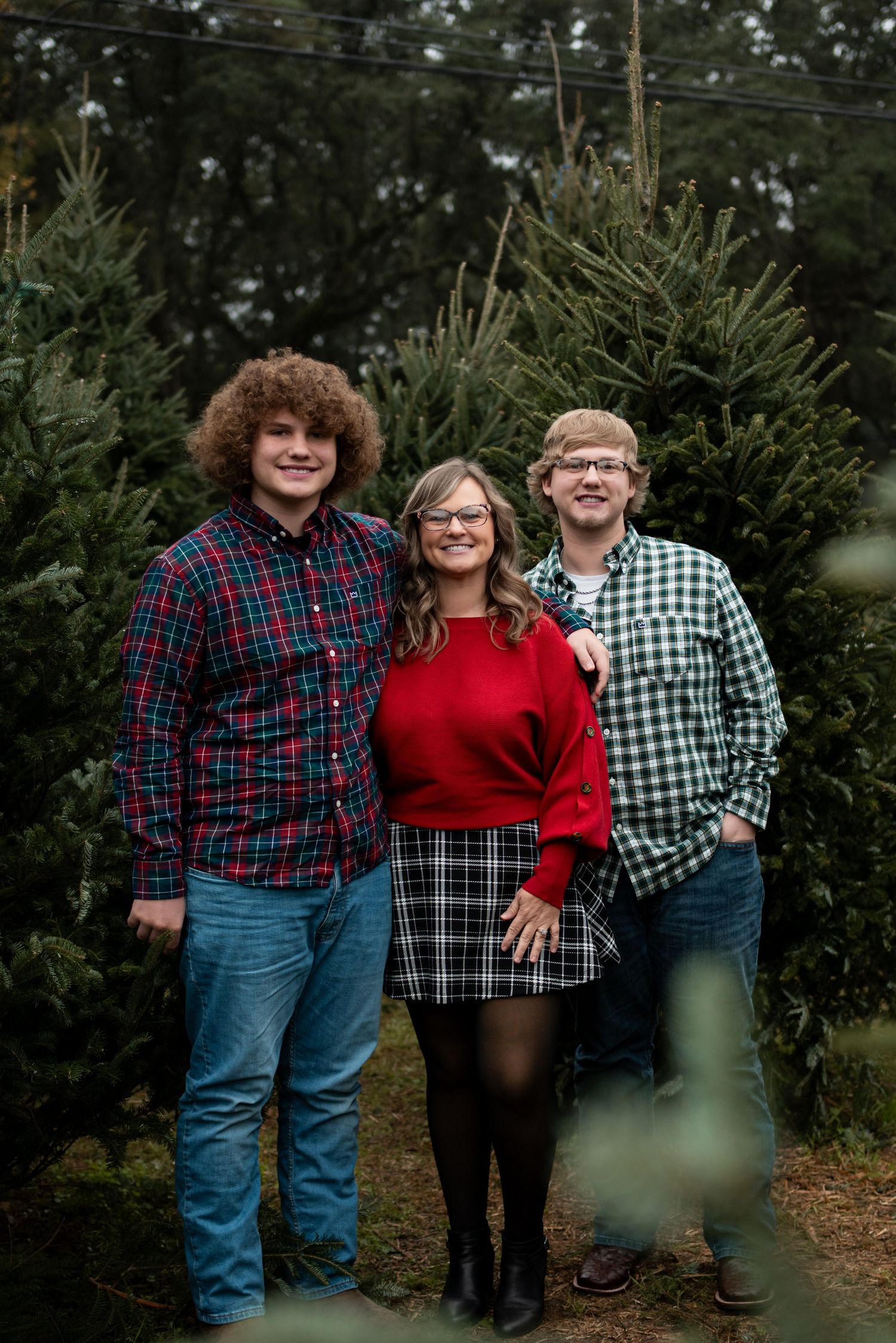 Family of three posing at a Christmas tree farm, wearing festive plaid outfits, smiling among lush green pine trees.