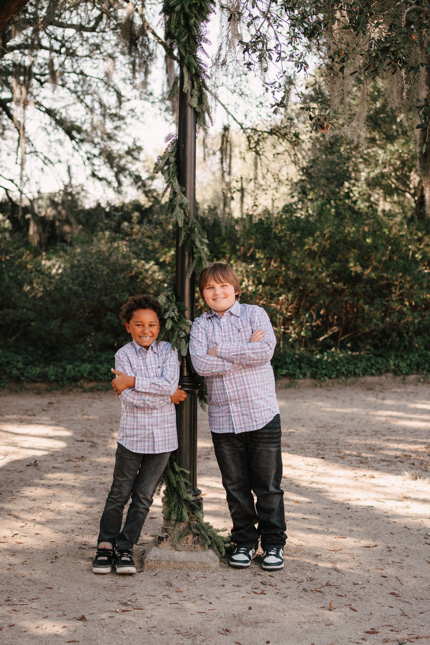 Two boys in matching plaid shirts stand arms crossed by a decorated pole outdoors among mossy trees.
