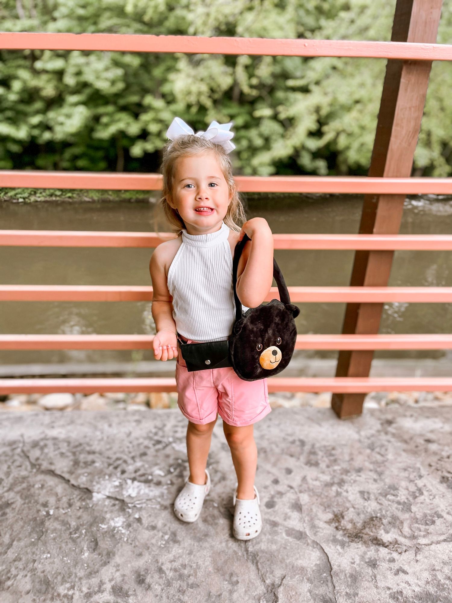 A cute toddler girl with a white bow poses on a bridge wearing a white top, pink shorts, and a bear purse.