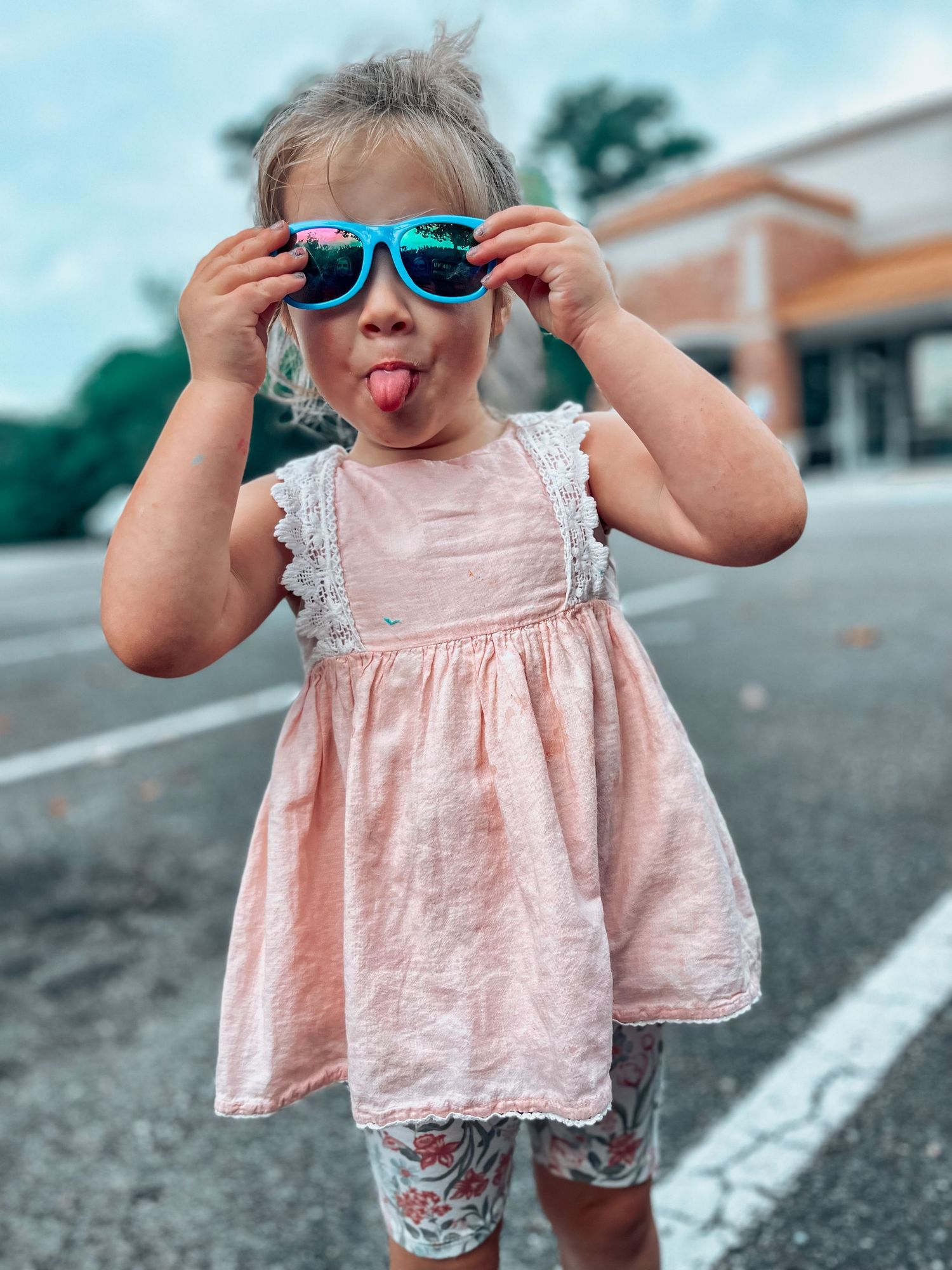 A playful toddler girl sticks out her tongue while holding blue sunglasses in a parking lot setting.