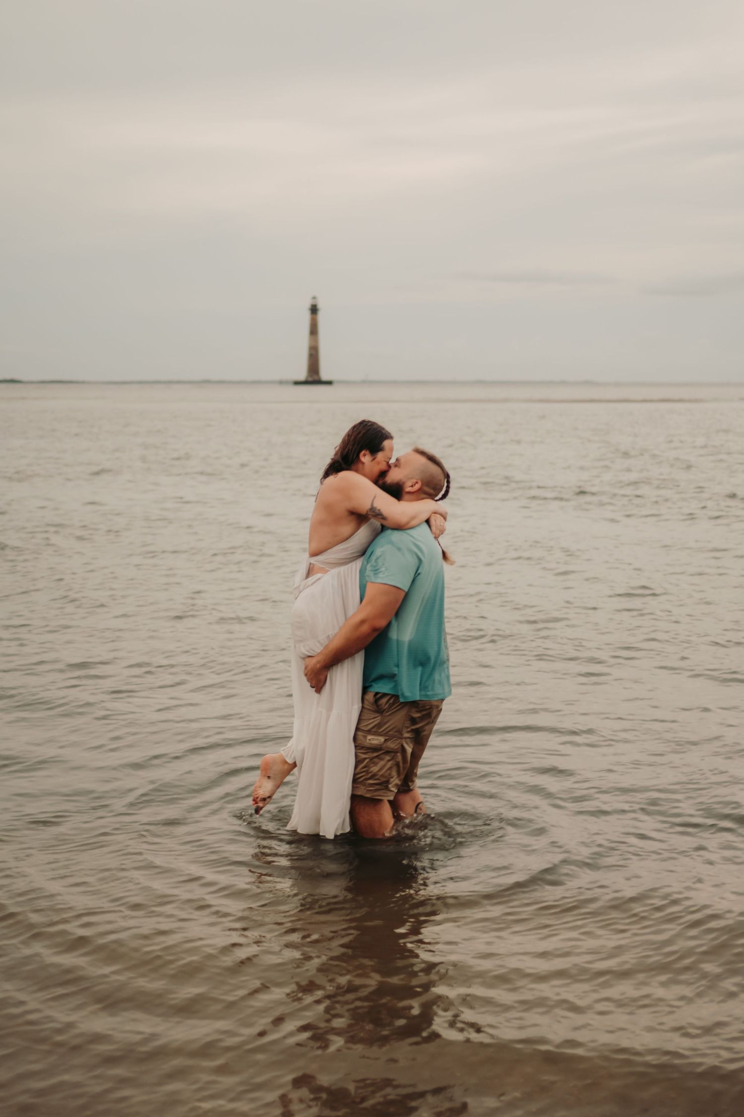 Couple kissing in shallow ocean water with a lighthouse in the background on a cloudy day.