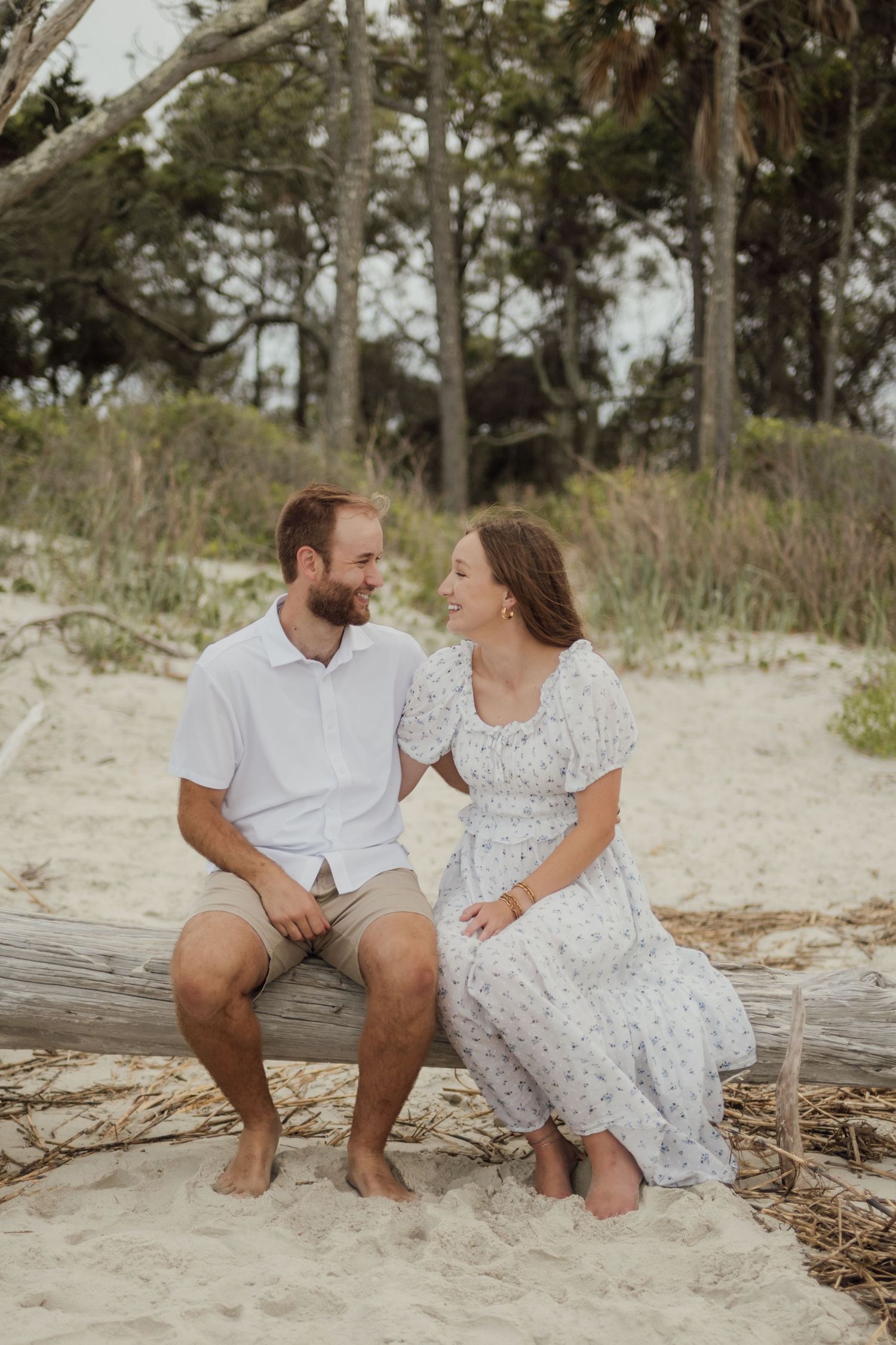 A couple sits on driftwood at a sandy beach, smiling at each other, dressed in casual white and floral outfits.
