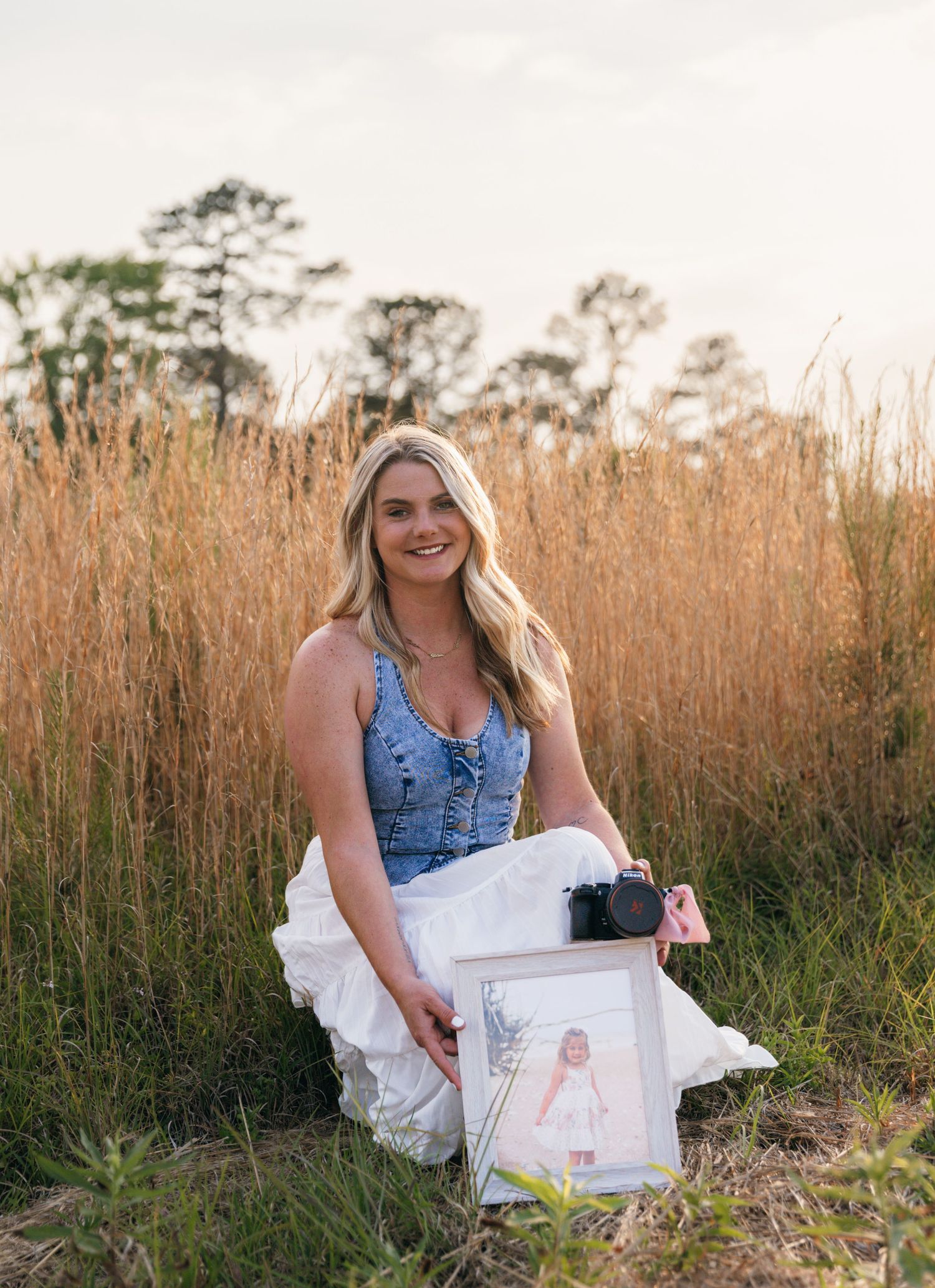 Blonde woman in denim top and white skirt sitting in golden field holding a framed photo and camera.