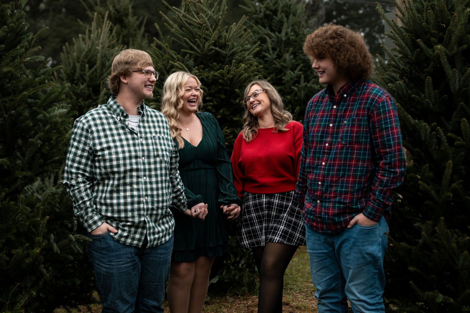 Four people laughing together at a Christmas tree farm, wearing festive plaid and holiday outfits.