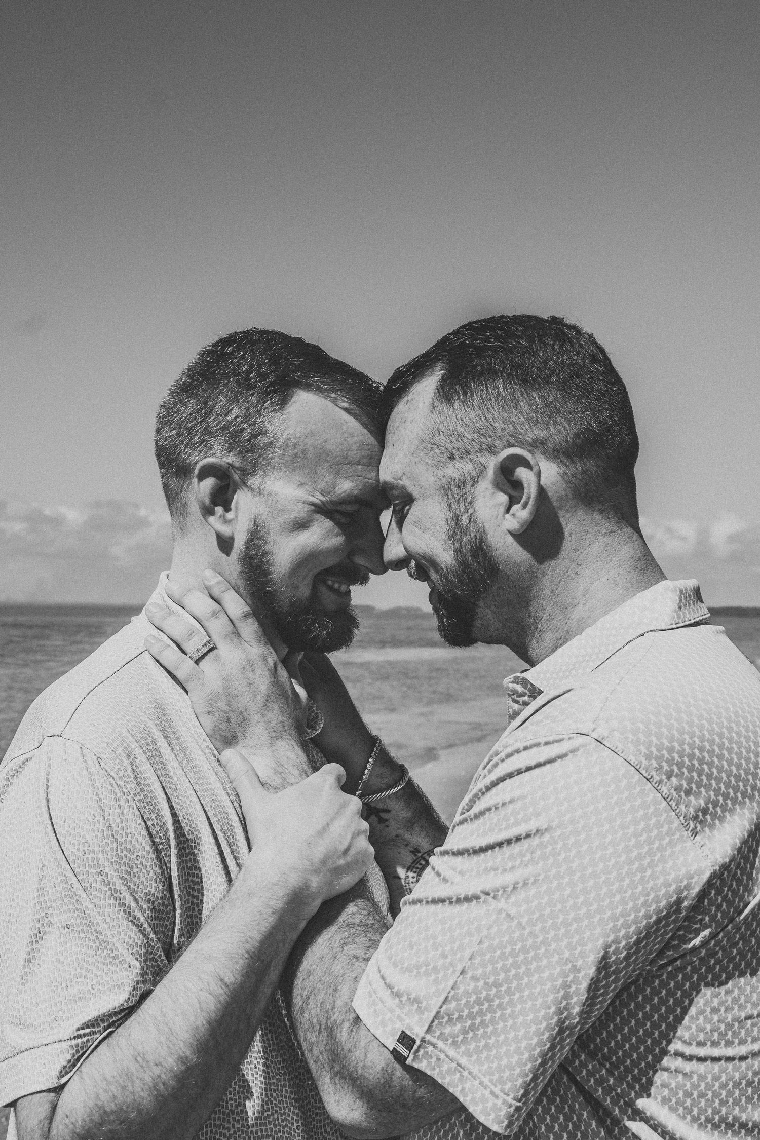 Black and white photo of two bearded men smiling and touching foreheads near the ocean.
