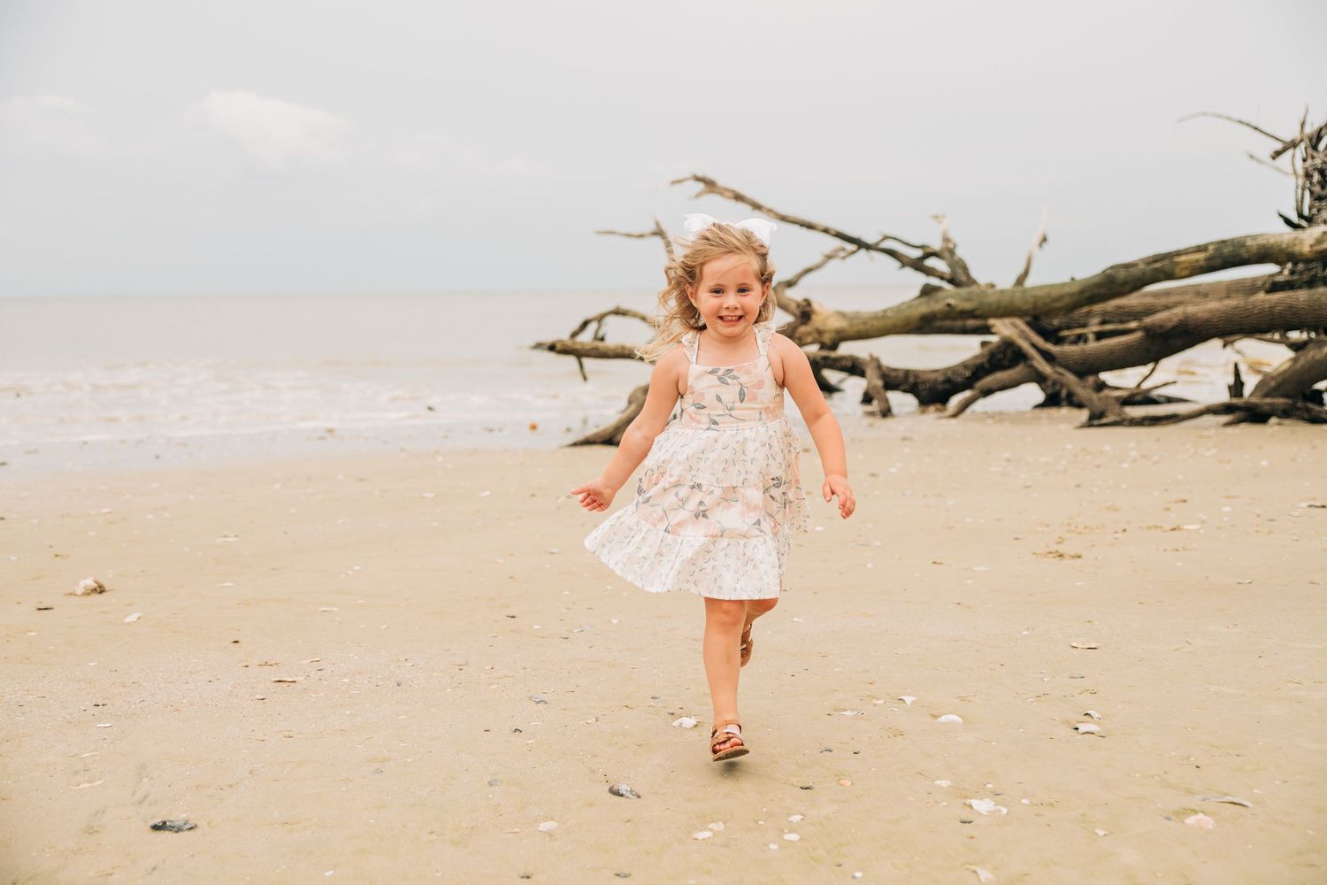 A happy little girl in a white dress runs along a sandy beach with driftwood and ocean in the background.