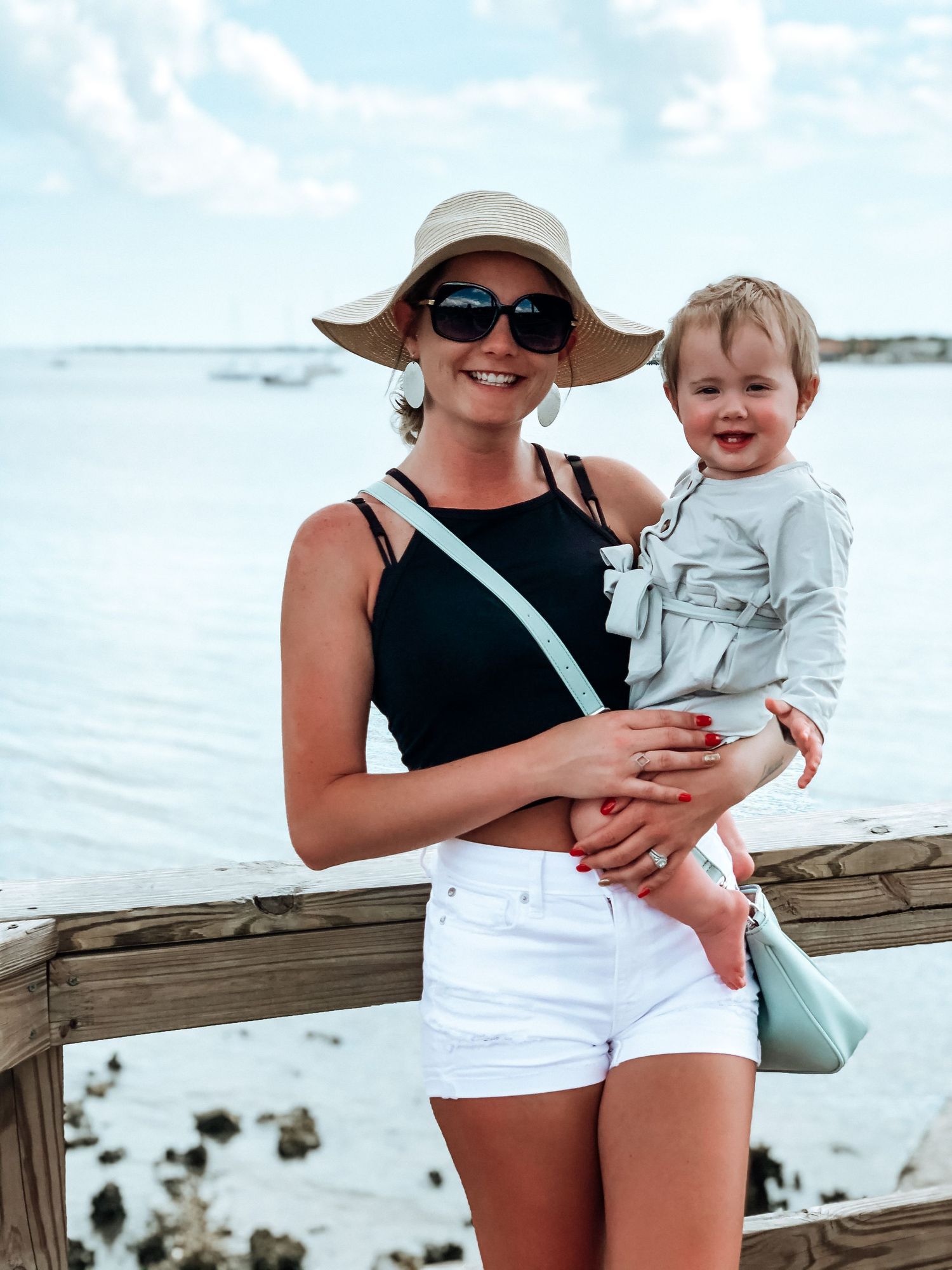 Smiling woman in sun hat and sunglasses holding toddler near waterfront on sunny day.