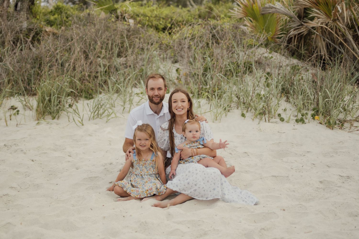 Family of four smiling together on sandy beach dunes, parents with two young children in light summer outfits.