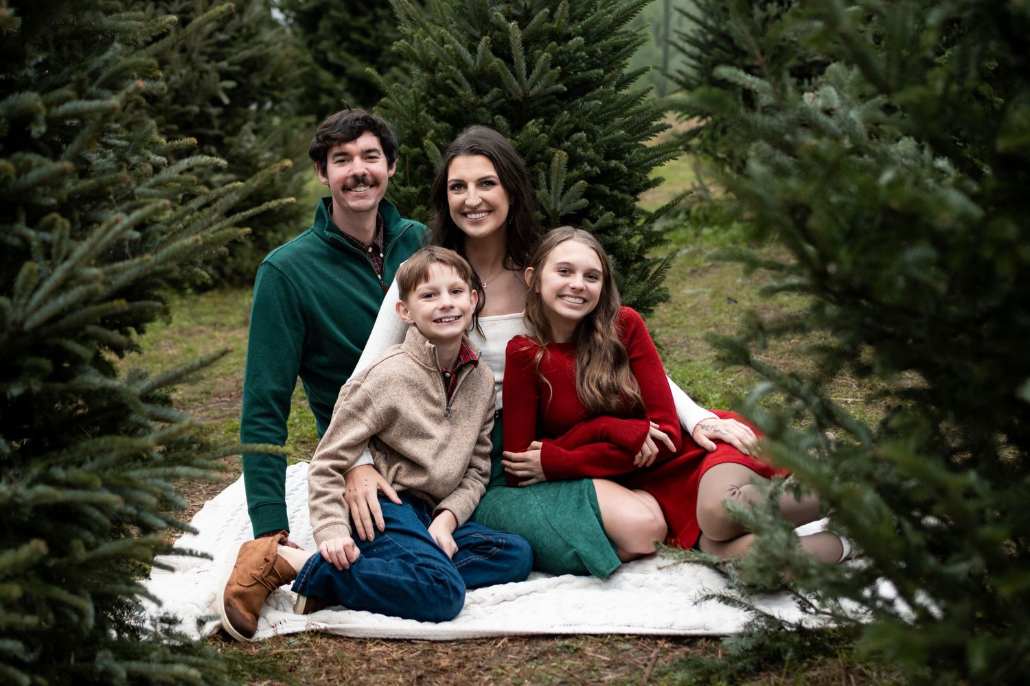 Family of four smiling together among Christmas trees at a tree farm during a holiday photo session.