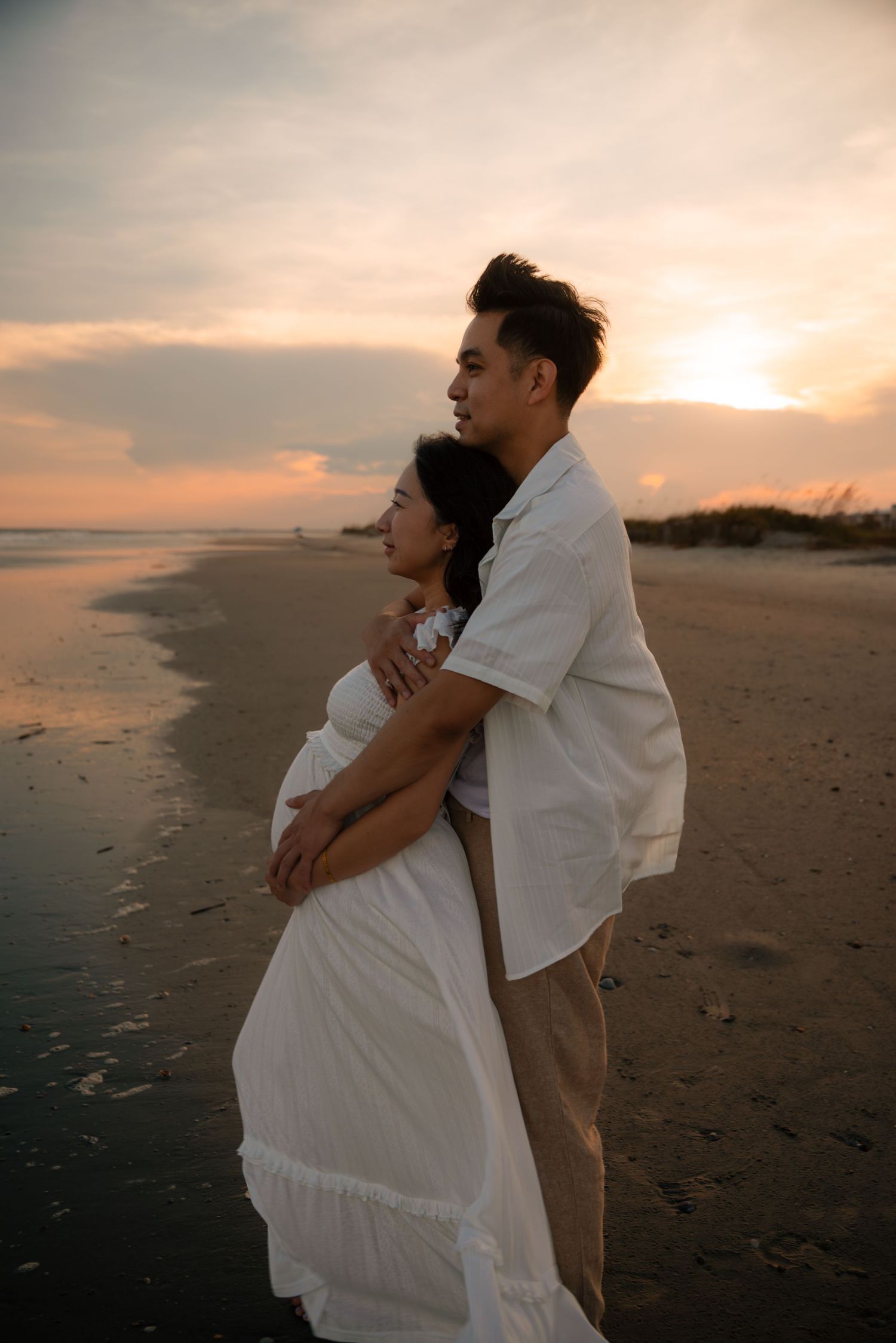 A couple embraces on a beach at sunset, the woman in a white dress cradling her baby bump as they gaze at the horizon.