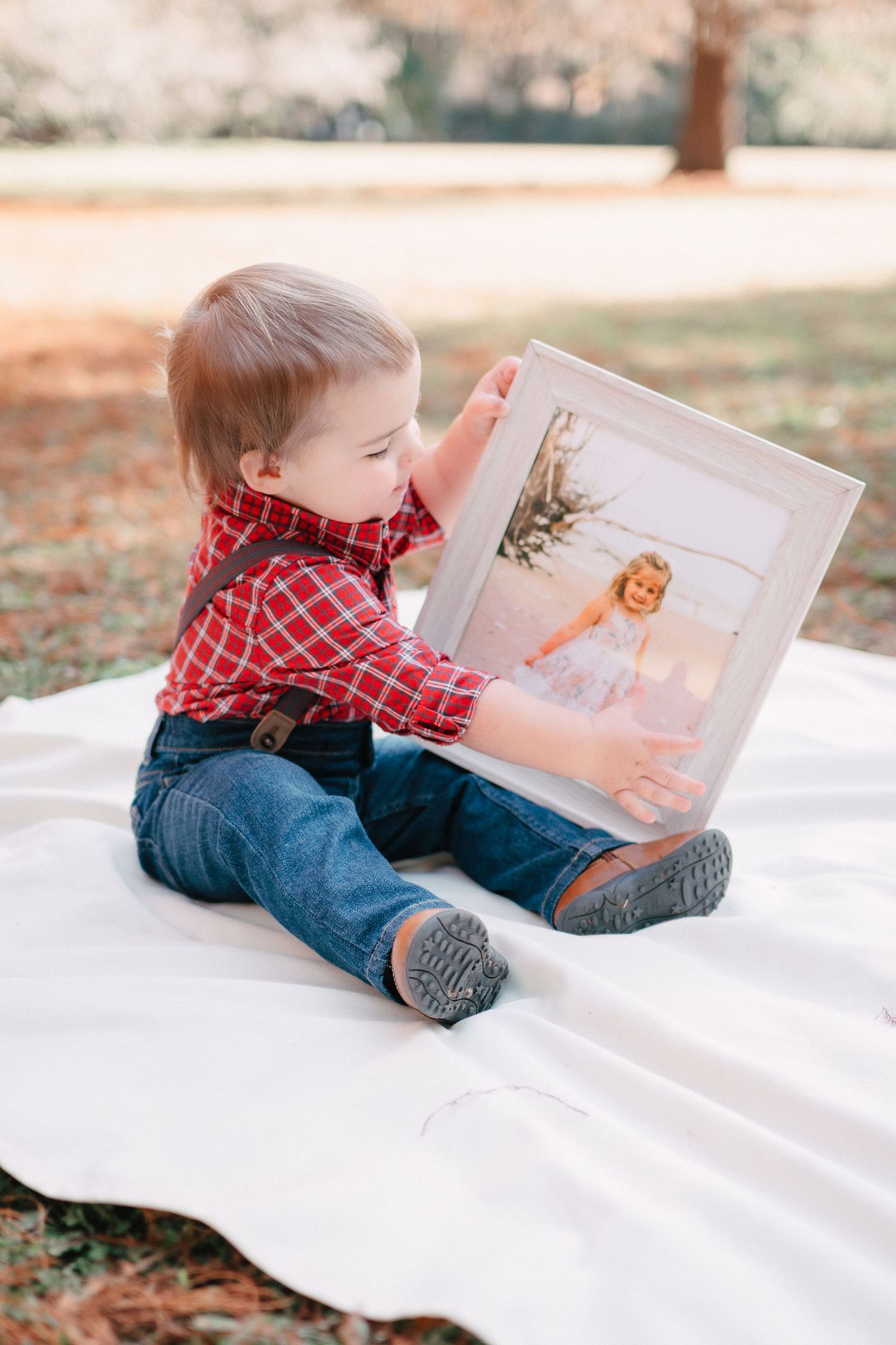 Baby boy in red plaid shirt sitting on white blanket outdoors holding a framed photograph.
