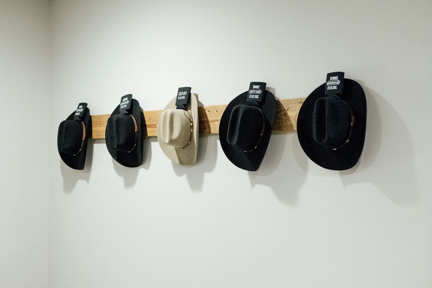 Five cowboy hats in black and tan mounted on a wooden rack against a white wall for a rustic wedding.
