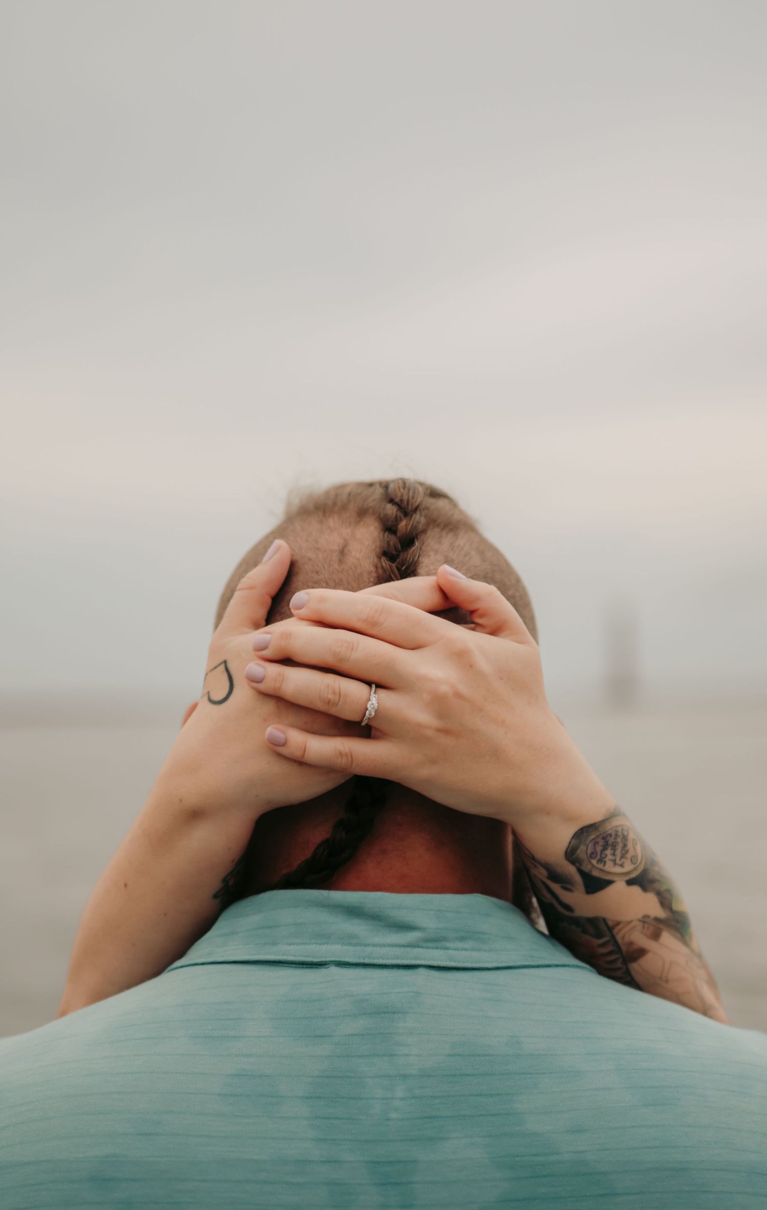 Person with tattoos and a ring clasping hands behind their head on a cloudy beach day.