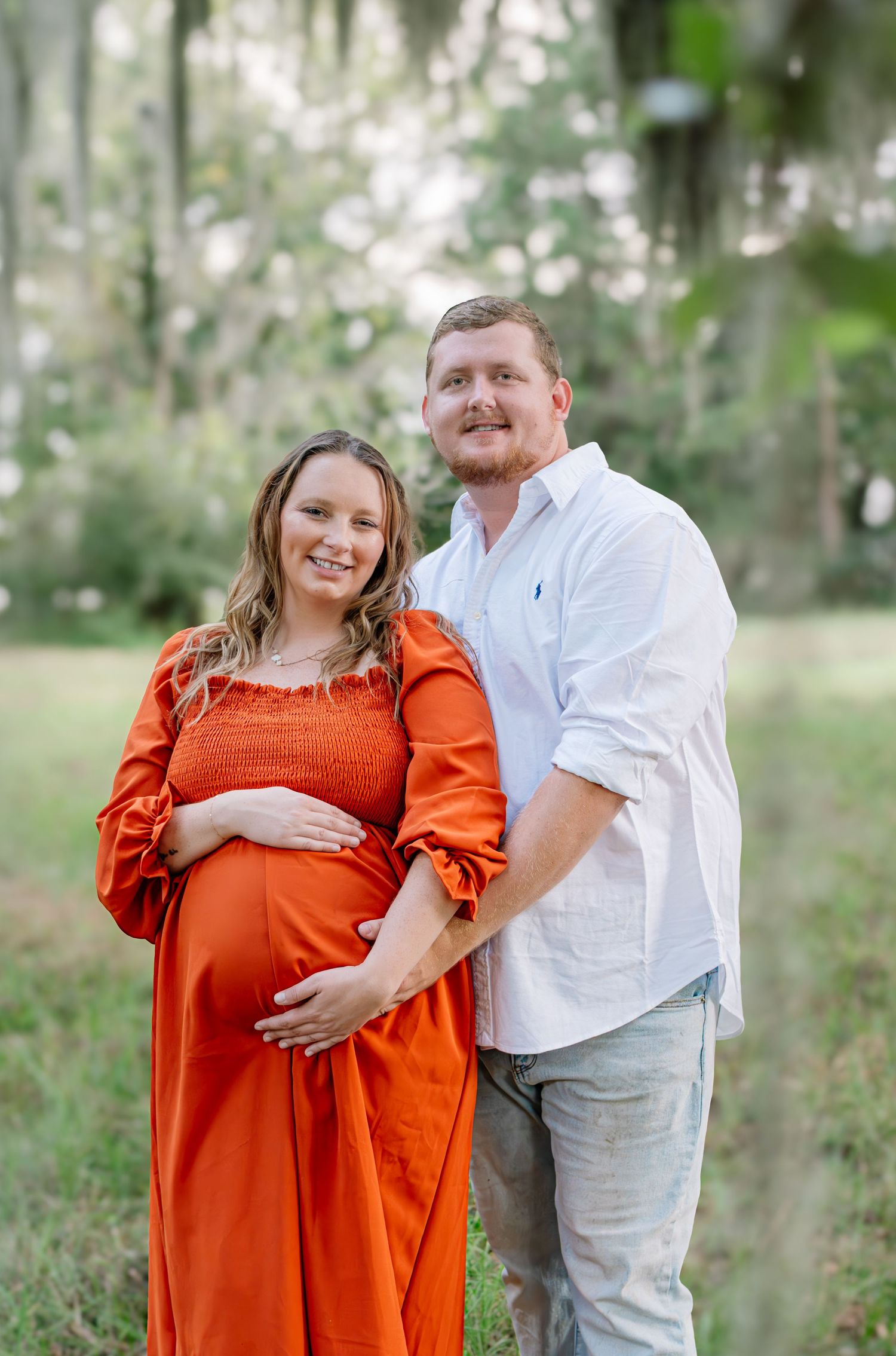 Pregnant woman in orange dress with partner outdoors during maternity photoshoot in lush green park.