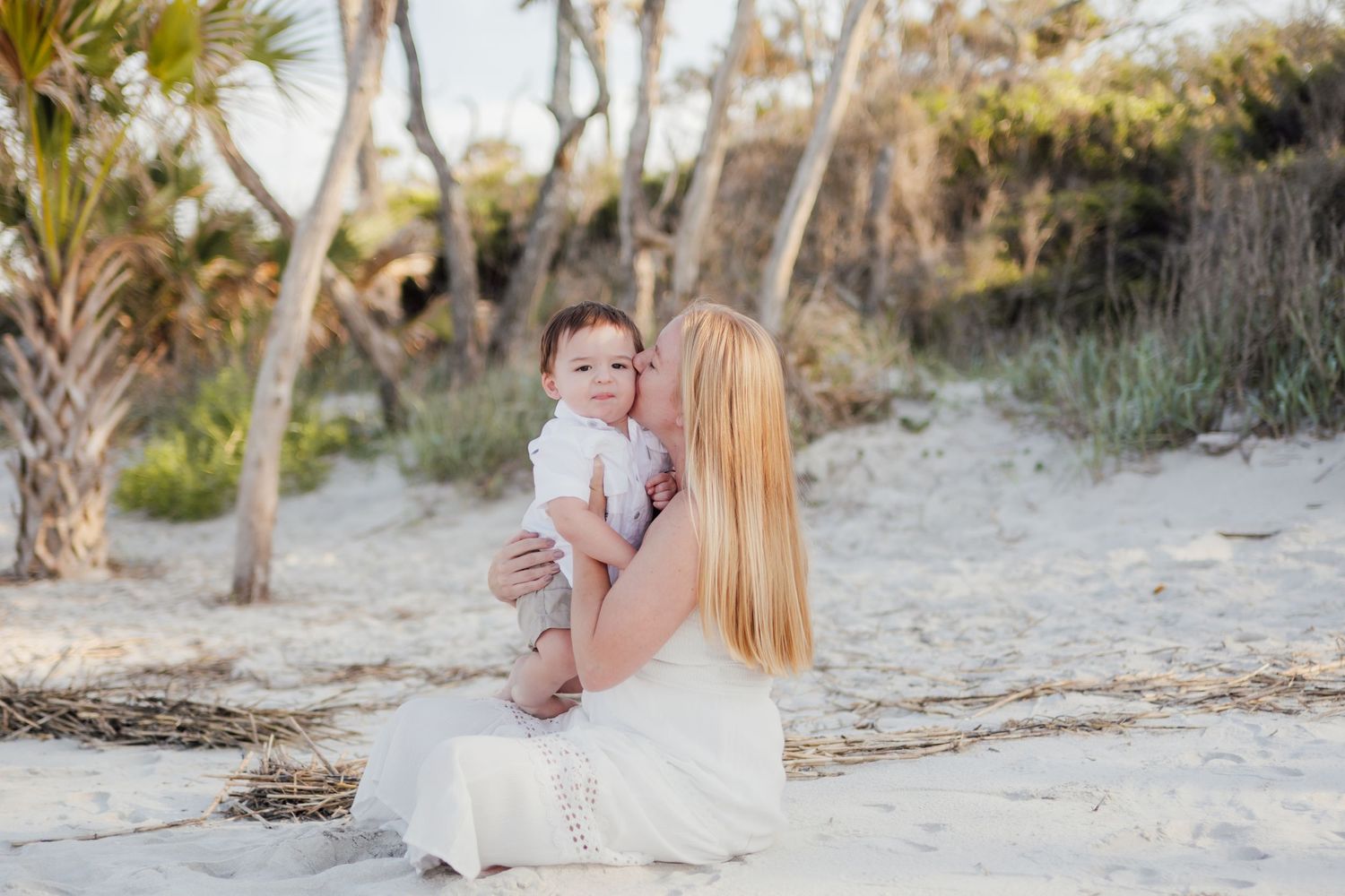 Mother in white dress kissing baby on beach with palm trees and sandy dunes in soft natural light.