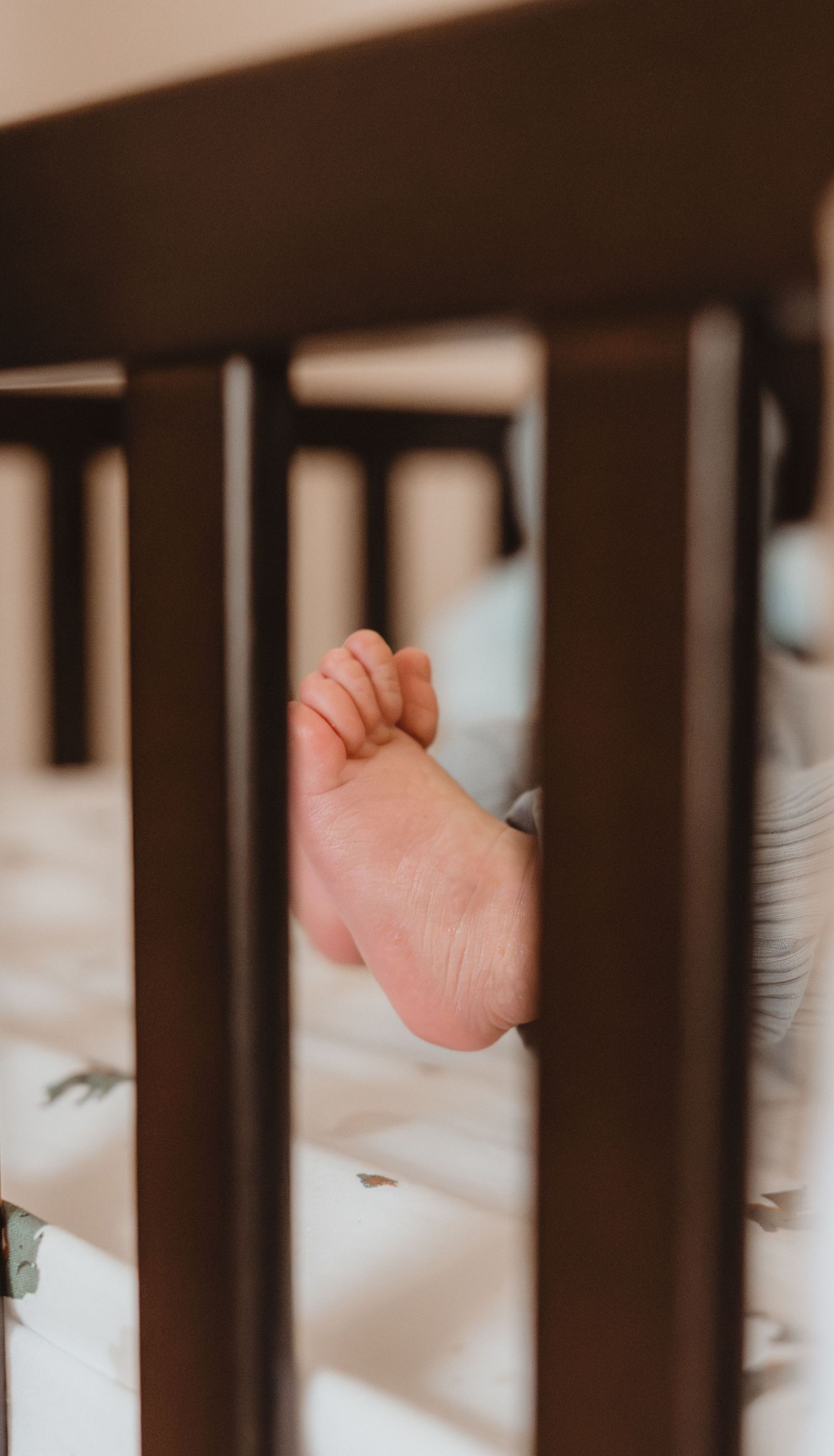 Close-up of a newborn baby's tiny foot peeking through dark wooden crib slats on a white patterned sheet.