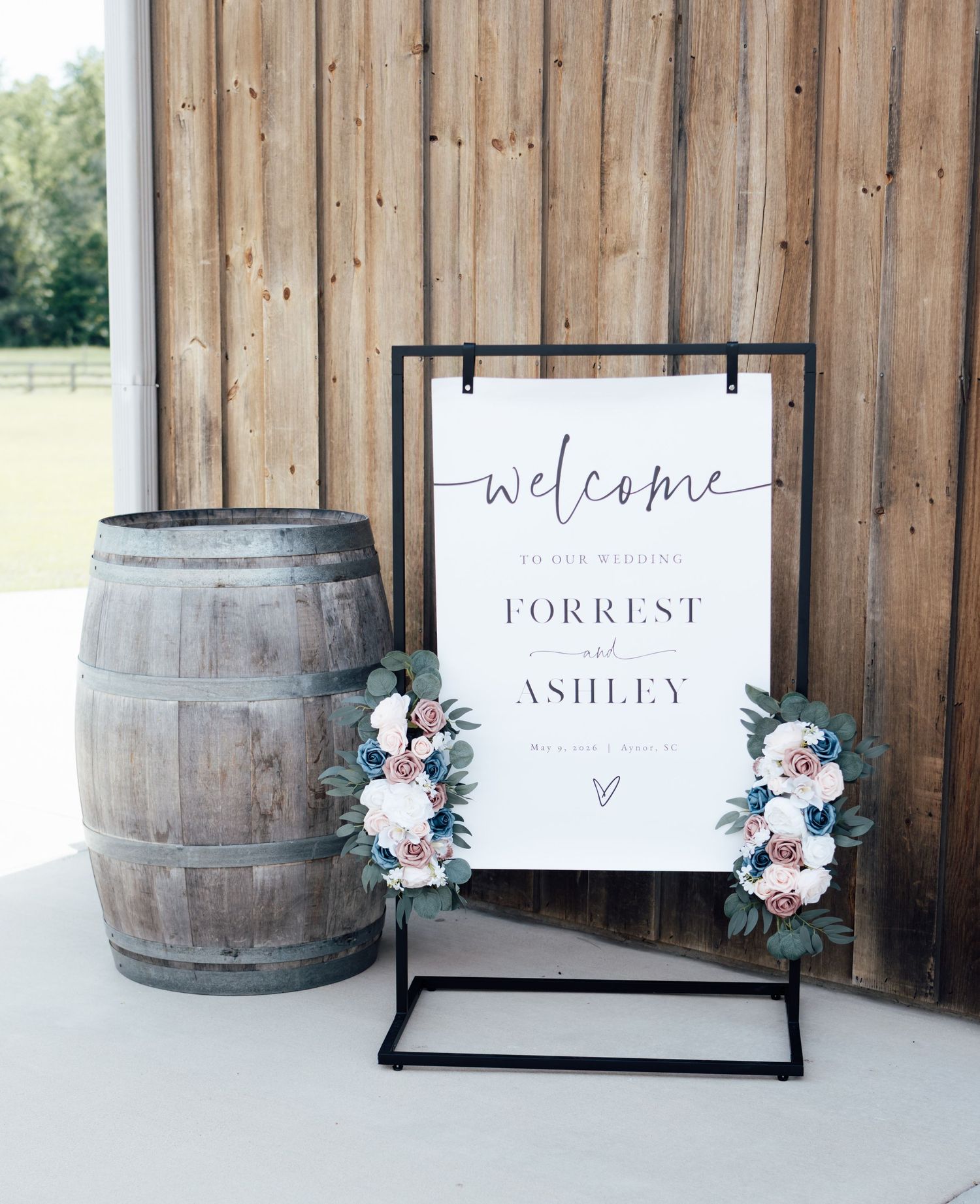 Elegant wedding welcome sign for Forrest and Ashley on black stand with floral accents beside a rustic wooden barrel.