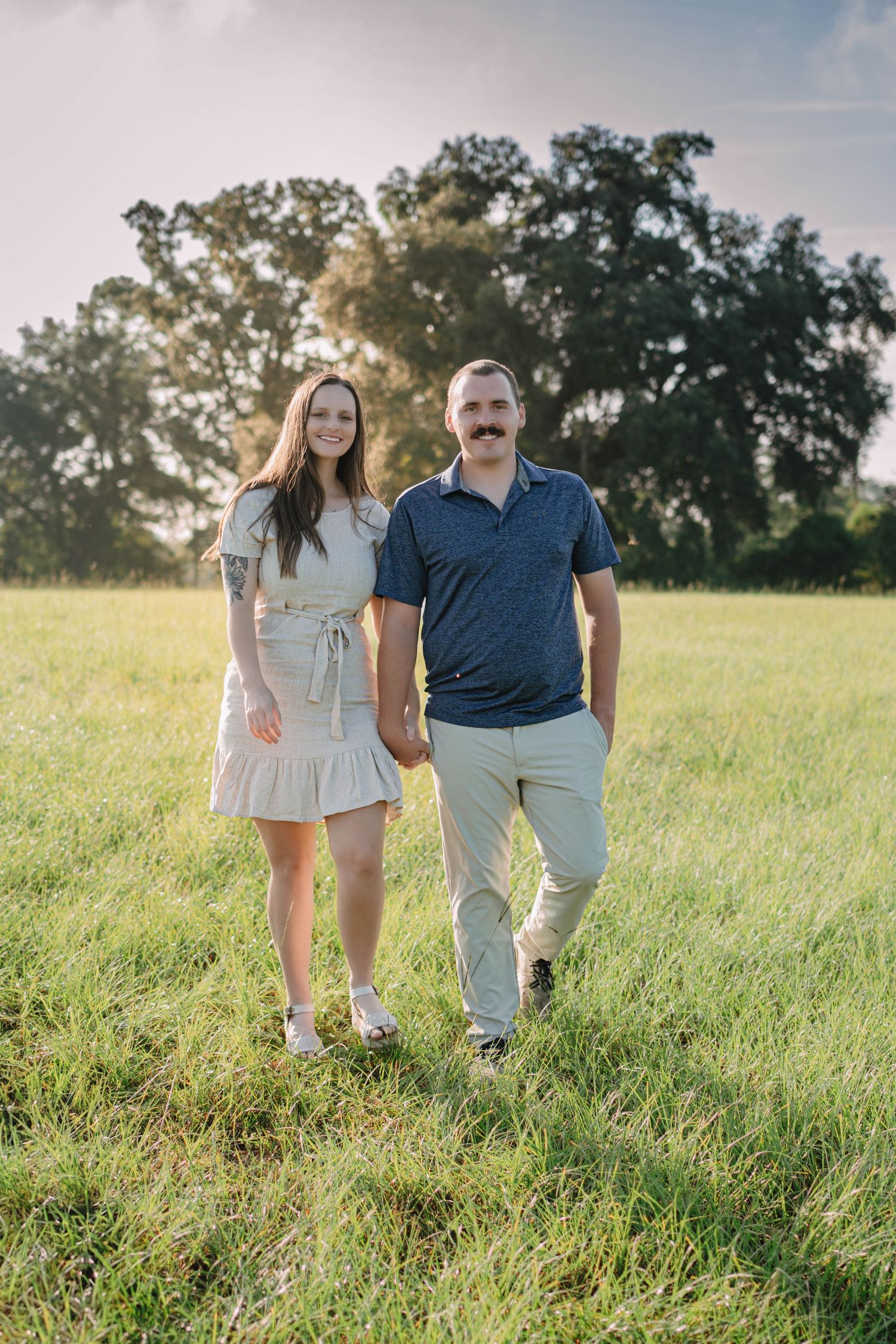 Couple holding hands smiling in a green field with large oak trees at golden hour.