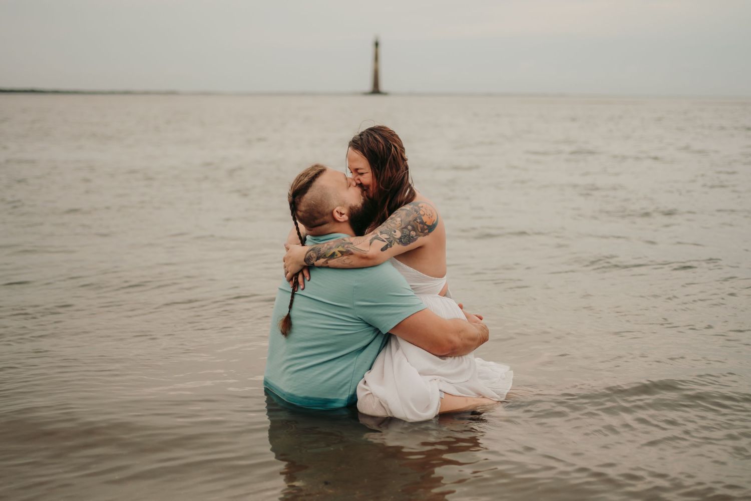Couple sharing a romantic kiss while sitting in shallow ocean water near a lighthouse.
