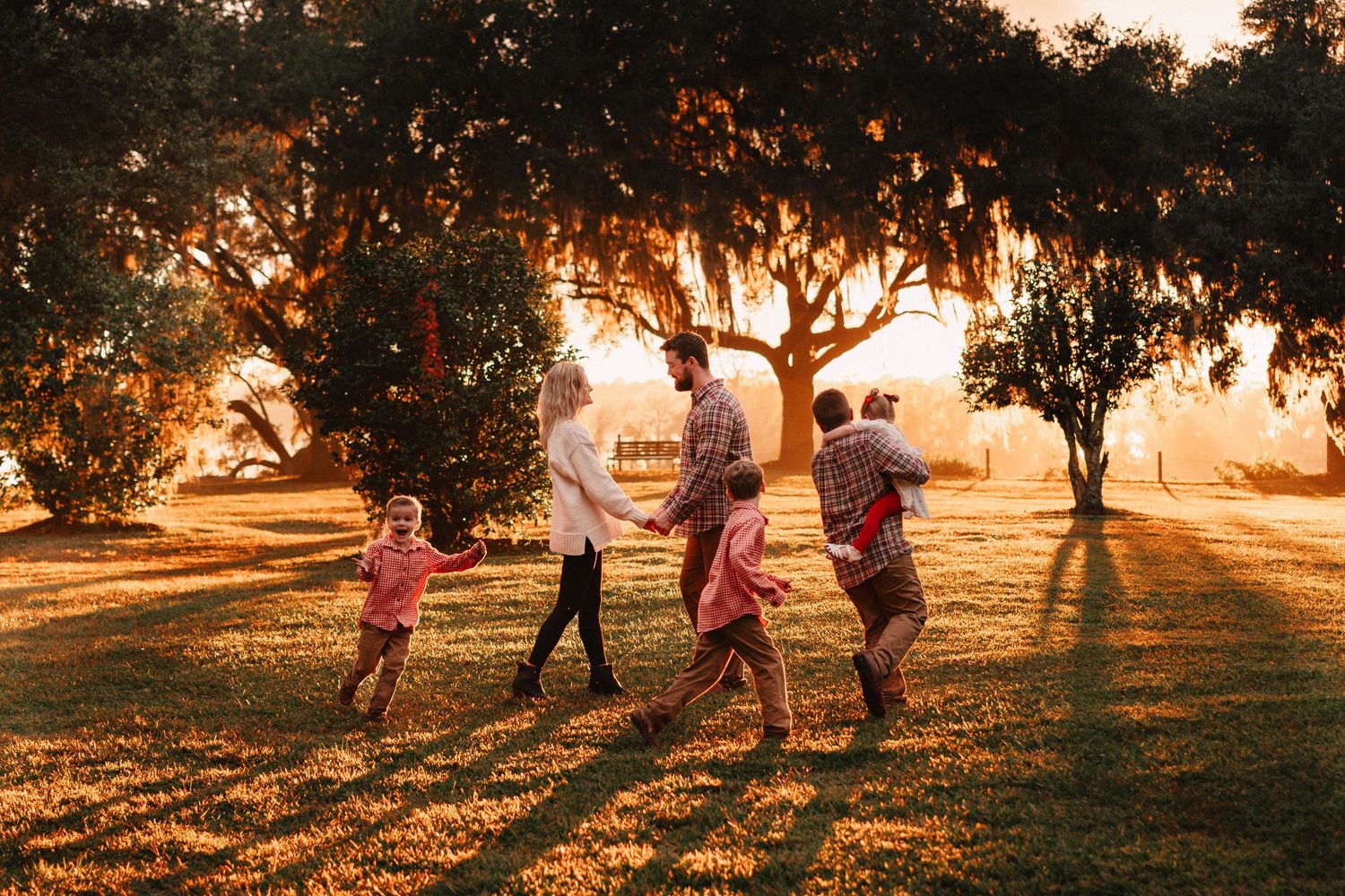 Family playing outdoors at golden hour under large oak trees on a sunny green lawn.