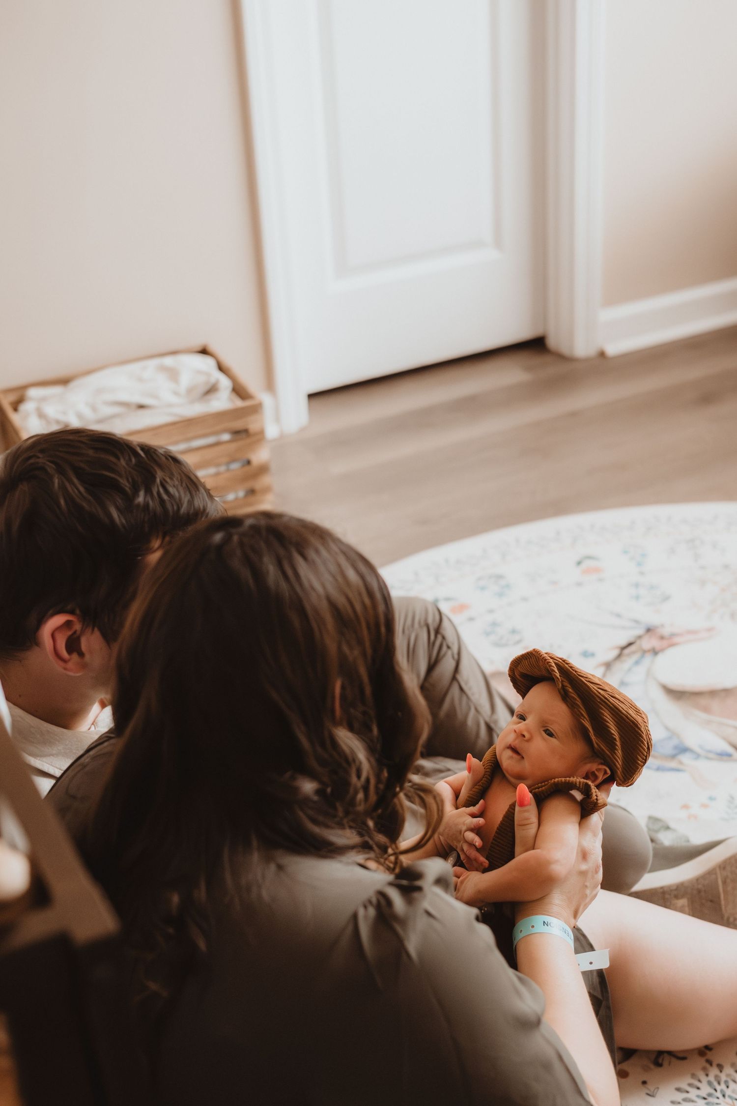 Parents sitting on floor bonding with newborn baby wearing brown hat in bright modern nursery room.