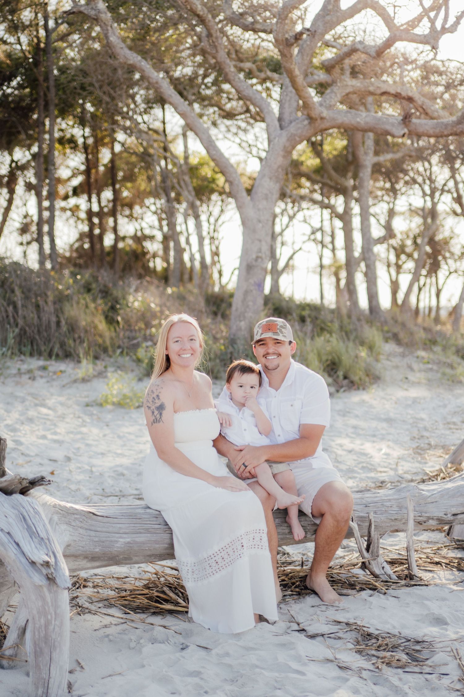 A smiling family of three poses on driftwood at a sandy beach, with a woman in white dress, man in cap, and baby.