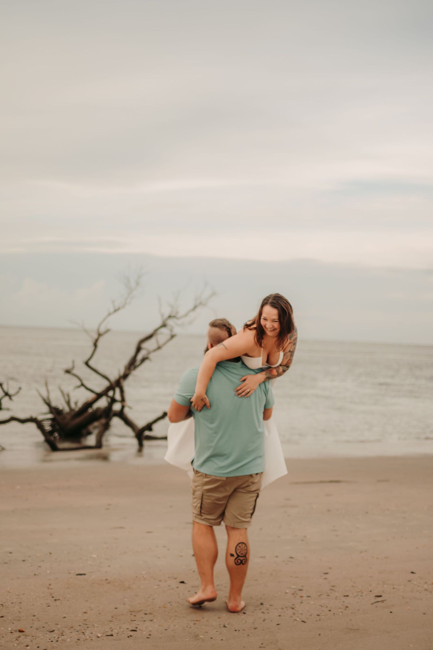 Couple embracing on a beach, man lifting woman in a white dress, with a driftwood tree and ocean in the background.
