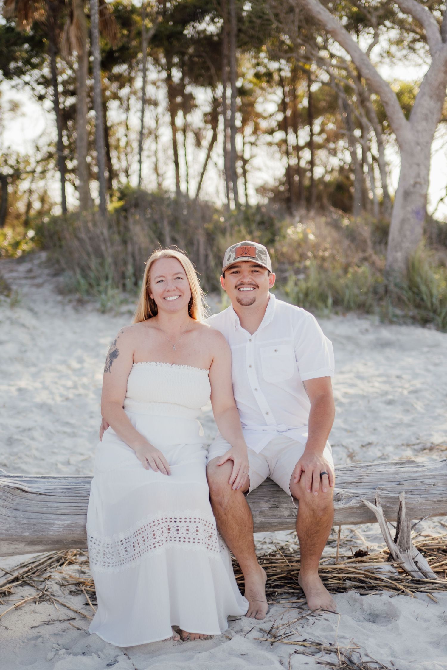 Couple in white outfits smiling on sandy beach with driftwood and trees in background.